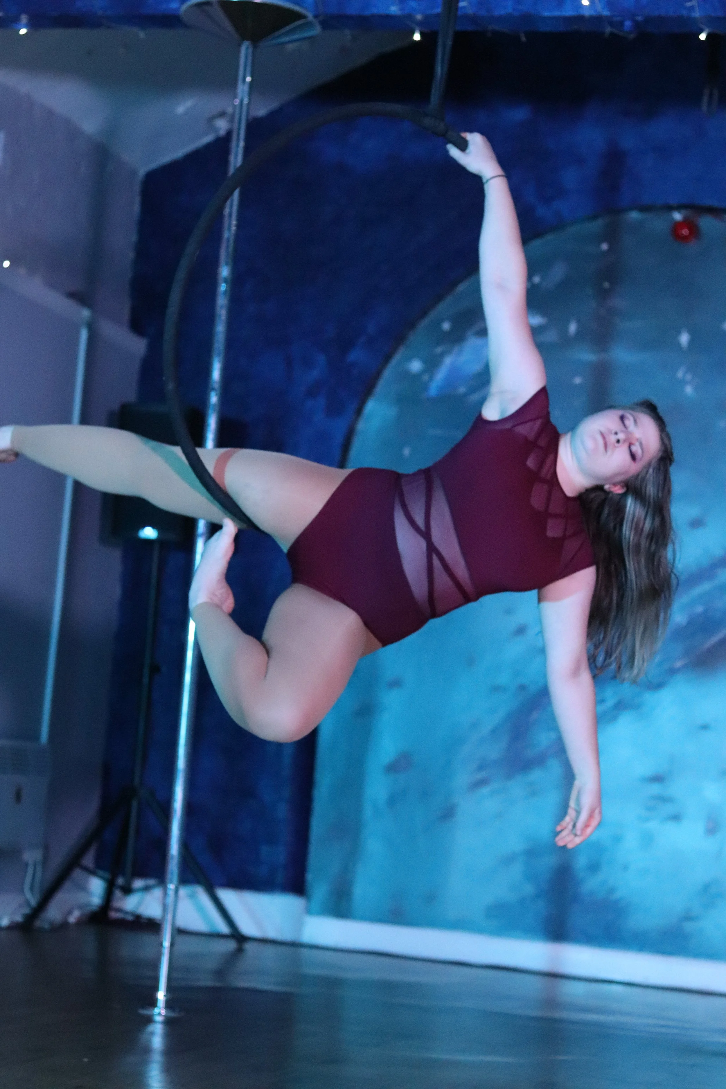 A woman performing aerial pole dance while suspended from a hoop, wearing a maroon outfit with sheer panels, against a dark blue background with a planetary design.