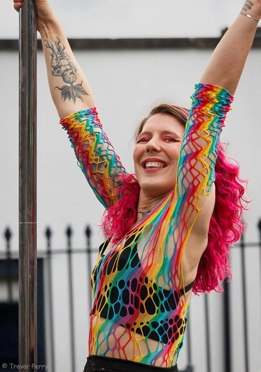Person with pink curly hair wearing a colorful rainbow fishnet dress, smiling and raising arms, holding a pole, with a tattoo on their arm and a fence in the background.