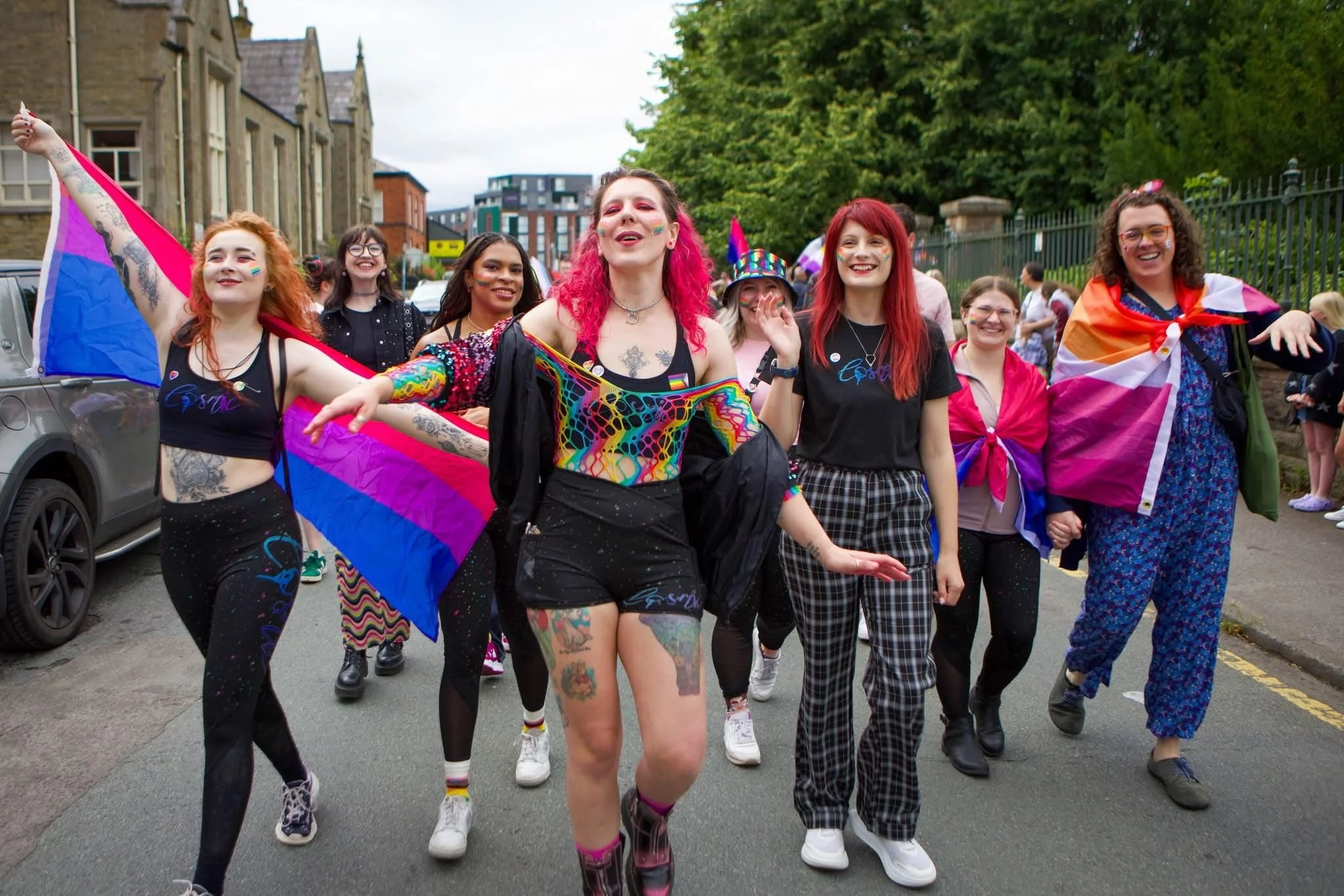 Group of people celebrating in Pride parade, some holding LGBTQ+ flags, dressed in colorful clothing and accessories.