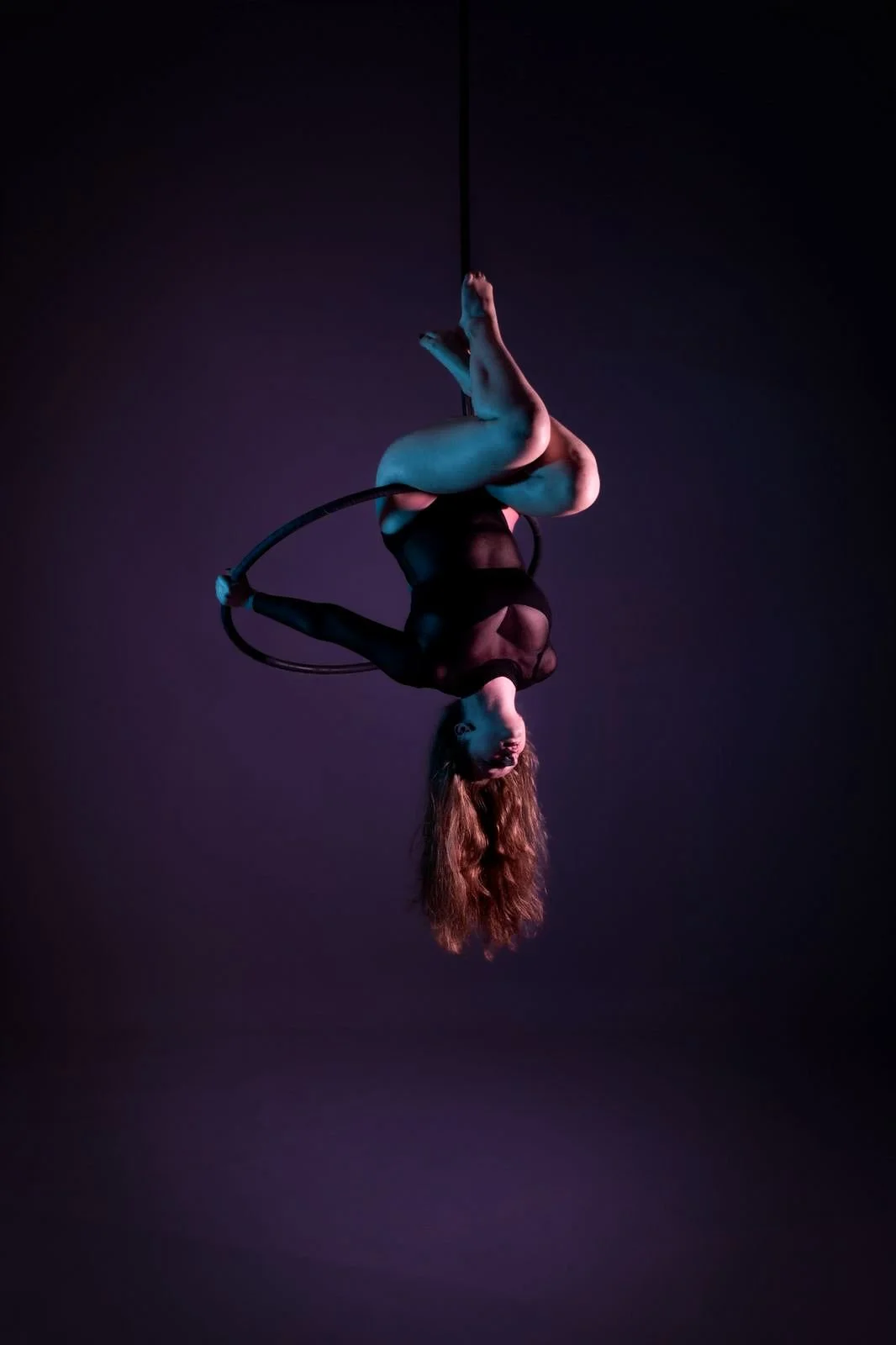 A woman performing aerial acrobatics on a hoop, hanging upside down in a dark studio with purple lighting.