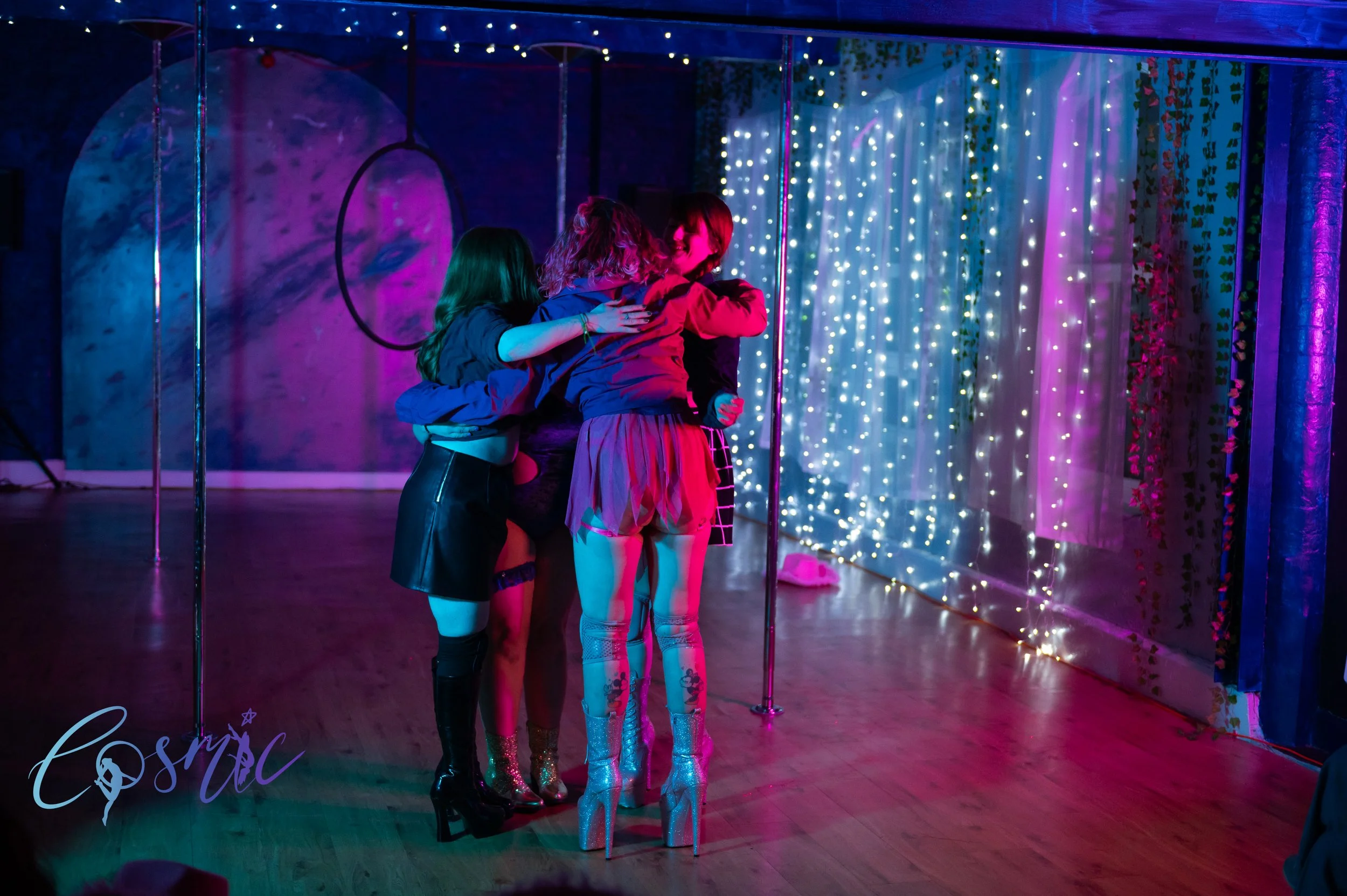 Four women in colorful clothing hugging each other on a dance floor with purple and blue lighting, with a background of string lights and decorative curtains.