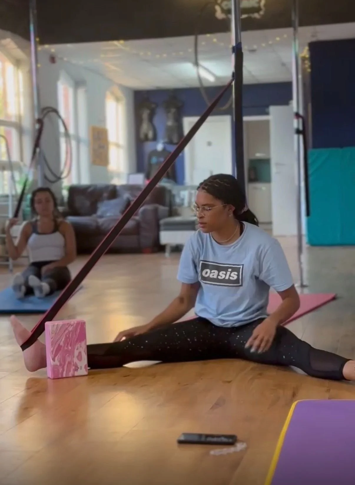A person performing a split stretch on the floor with a resistance band attached to a pink block, in a room with another person sitting and sitting with legs crossed in the background.