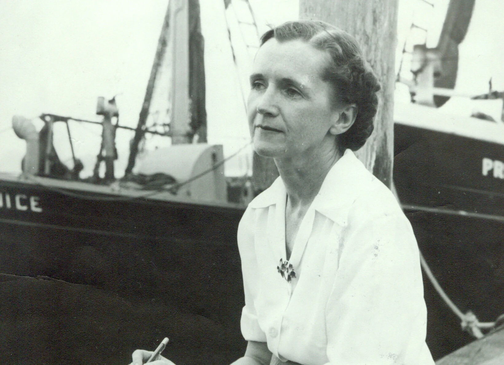 Black and white image of Rachel Carson sitting on a dock, looking pensively into the distance. A ship is behind her.