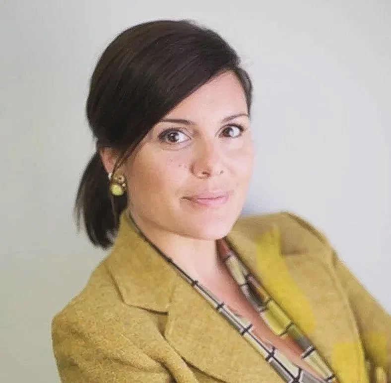 A woman with dark hair styled in a professional manner, wearing a mustard blazer, a geometric necklace, and matching earrings, against a neutral background.