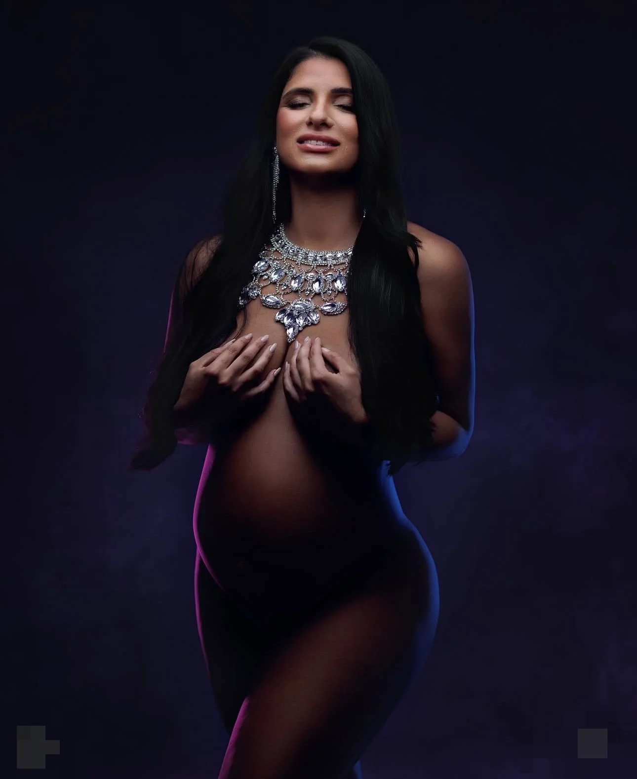 A woman in her maternity shoot with long black hair and jewelry covering her chest, posing topless against a dark background with colored lighting.