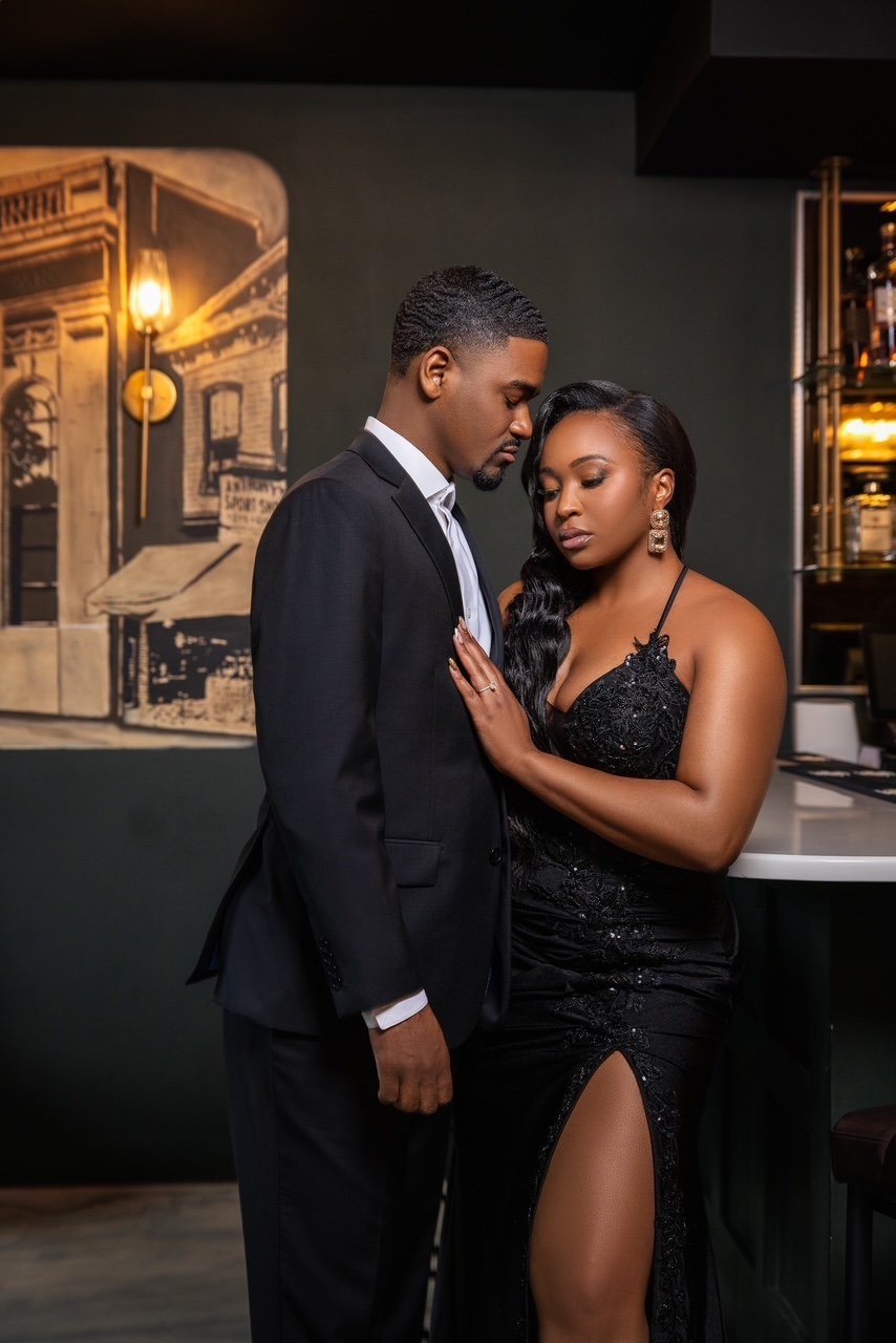 A man in a black suit and woman in a black dress with a high slit, standing close together in an intimate pose inside a dimly lit bar or restaurant.