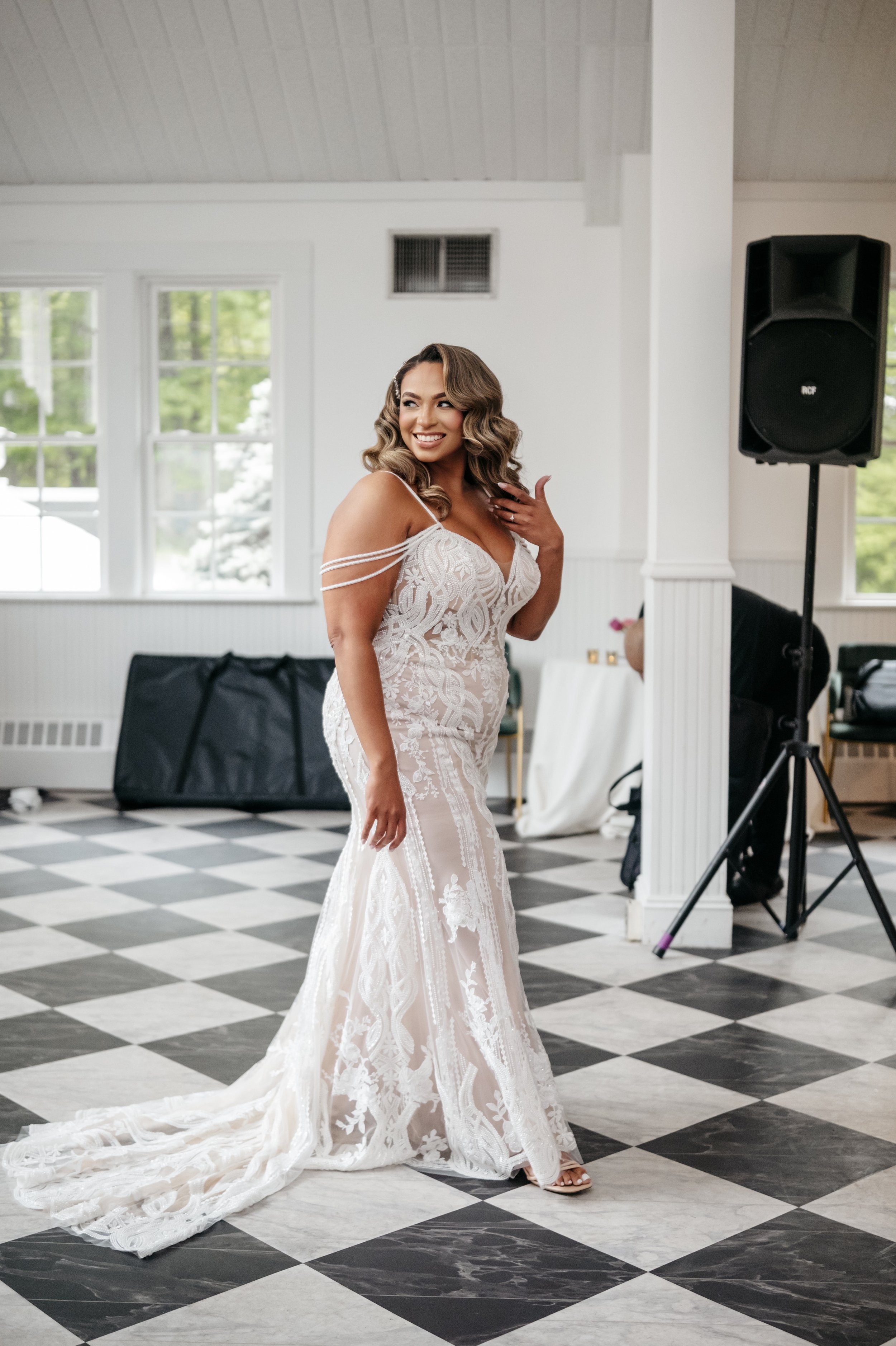 A woman in an elegant white lace wedding gown with off-the-shoulder straps stands on a black-and-white checkered floor, smiling and touching her hair inside a bright room with large windows and a speaker on a stand.