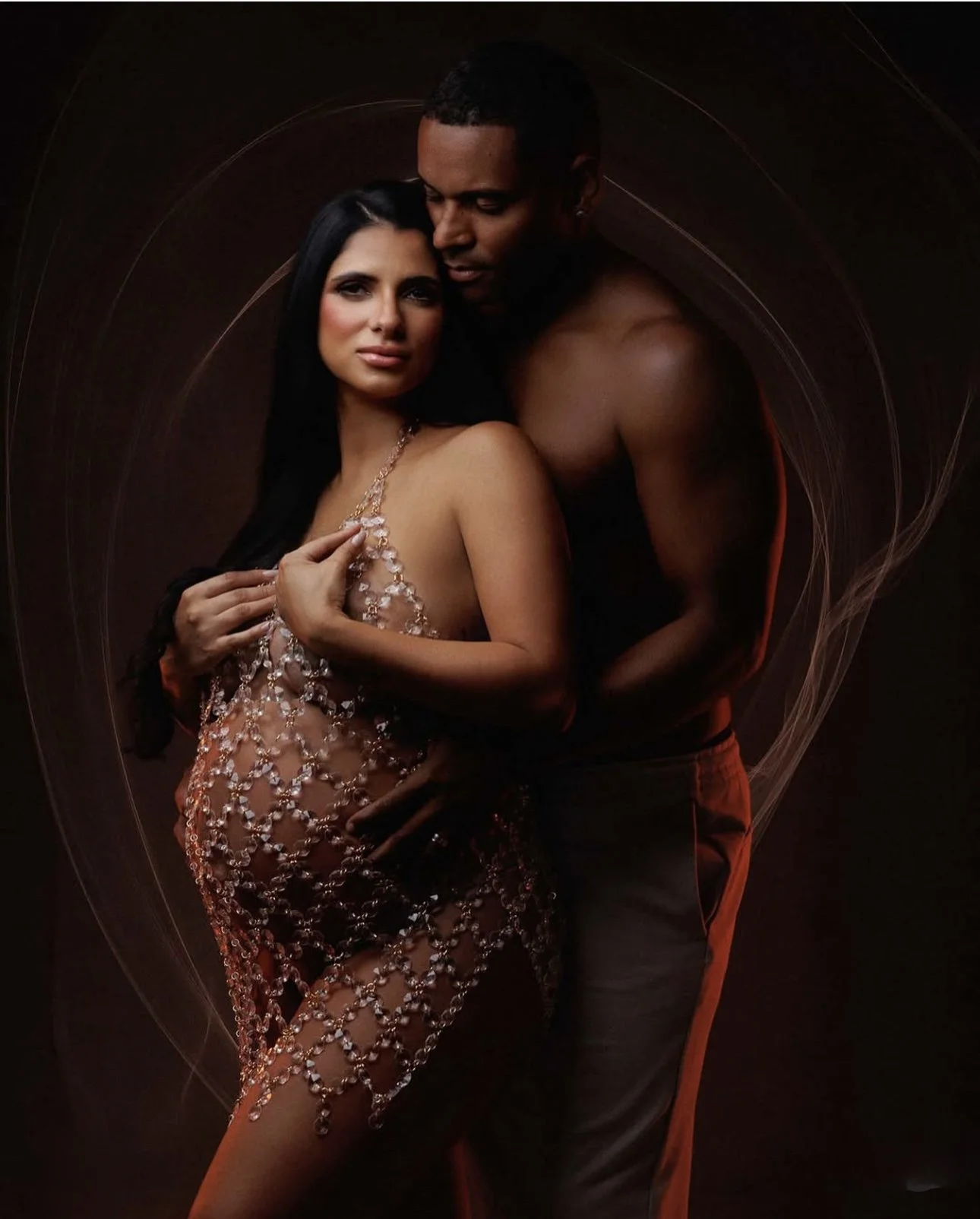 A woman at her maternity shoot with long dark hair and a man with short dark hair posing closely; she wears a revealing dress adorned with crystals, and he is shirtless.