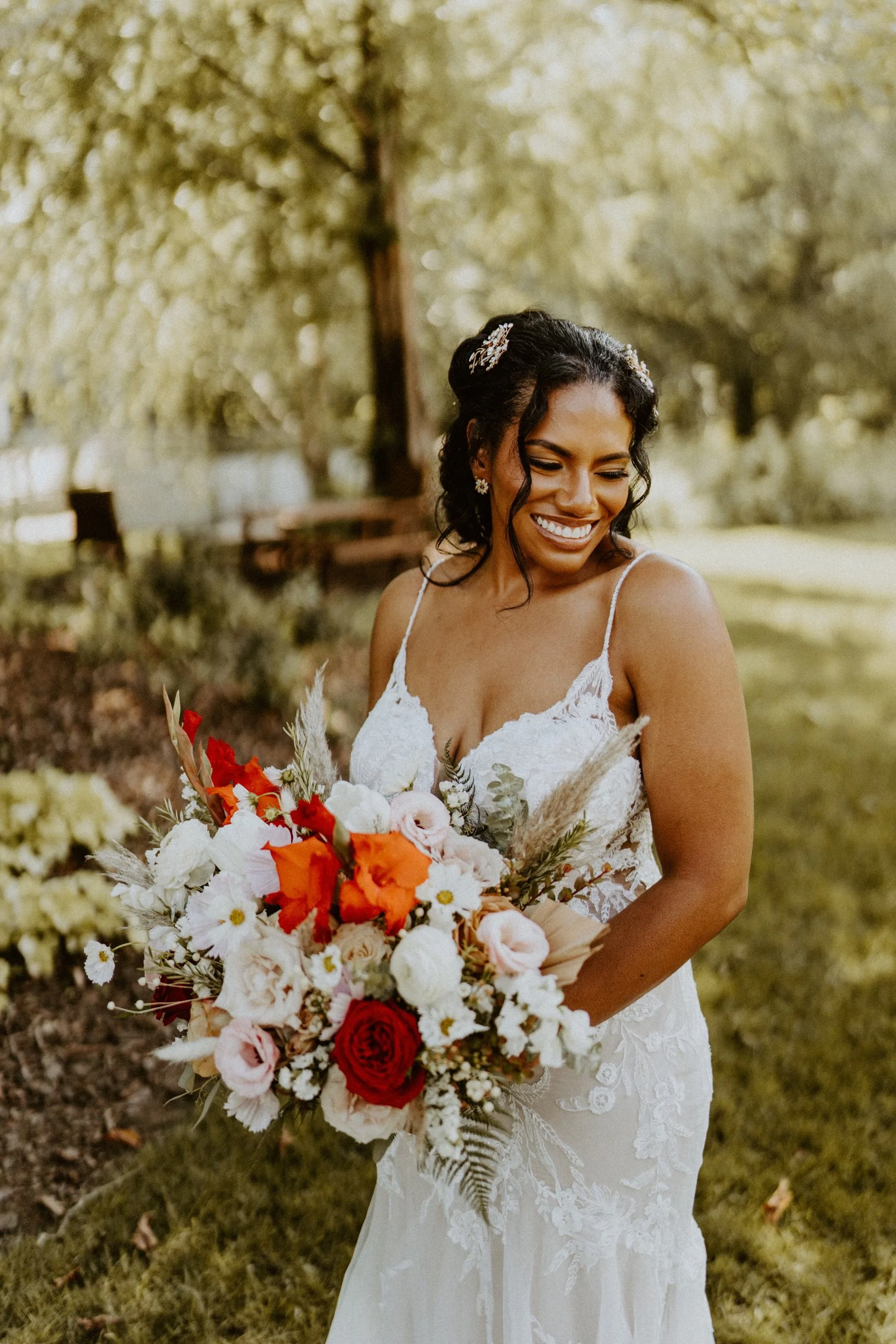 A bride outdoors holding a bouquet of colorful flowers, smiling and looking down, with a background of trees and greenery.