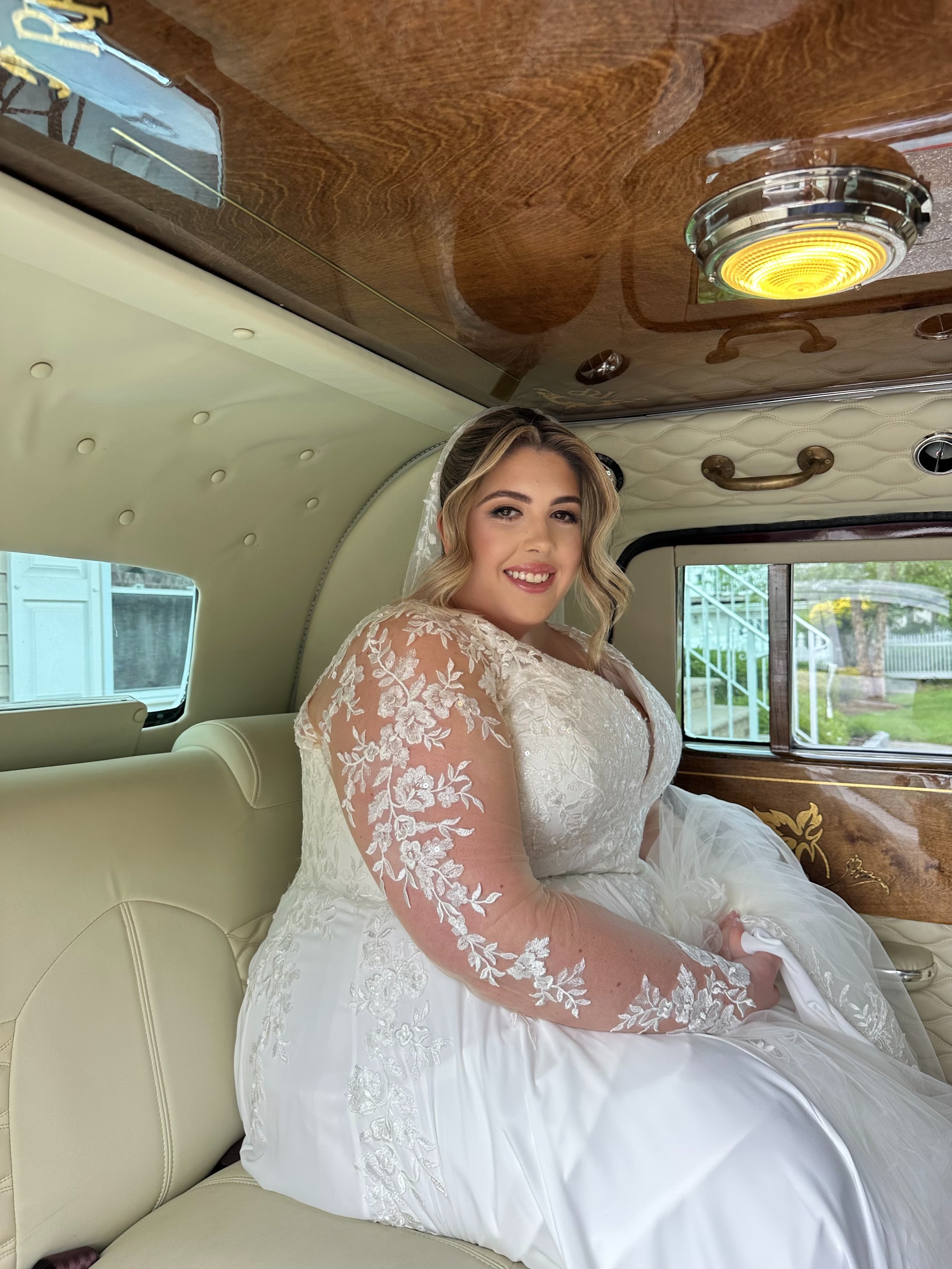 Bride in a white wedding dress with lace sleeves sitting in a vintage car with a cream interior and wooden paneling, smiling at the camera.