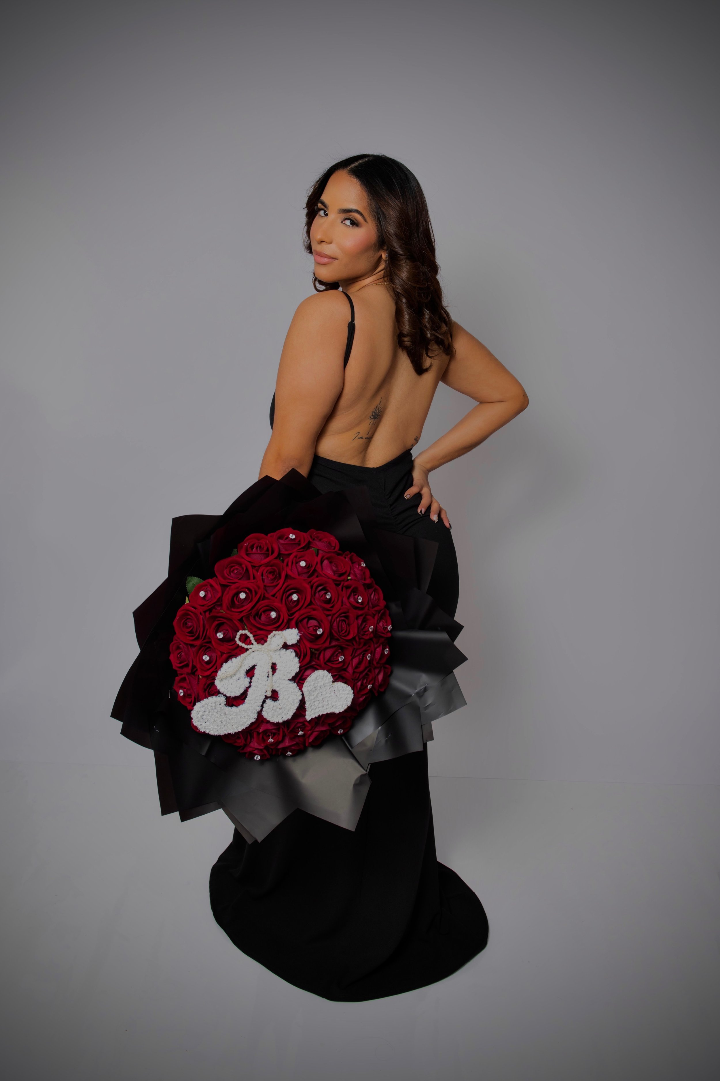 A woman in black attire holding a bouquet of red roses with a white 'R' and heart decoration.