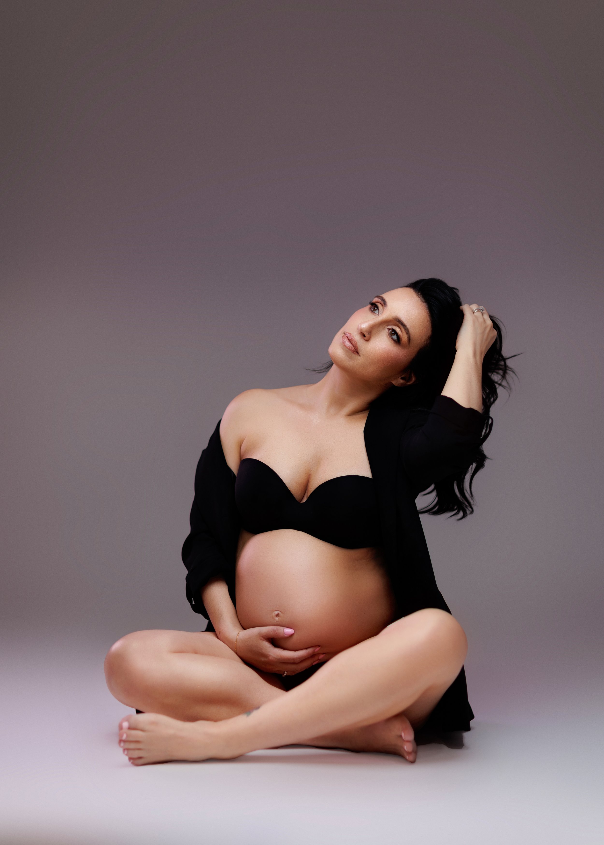 A pregnant woman sitting cross-legged on the floor, wearing a black bra and an open black shirt, with her hand on her belly and her other hand running through her hair against a neutral gray background.