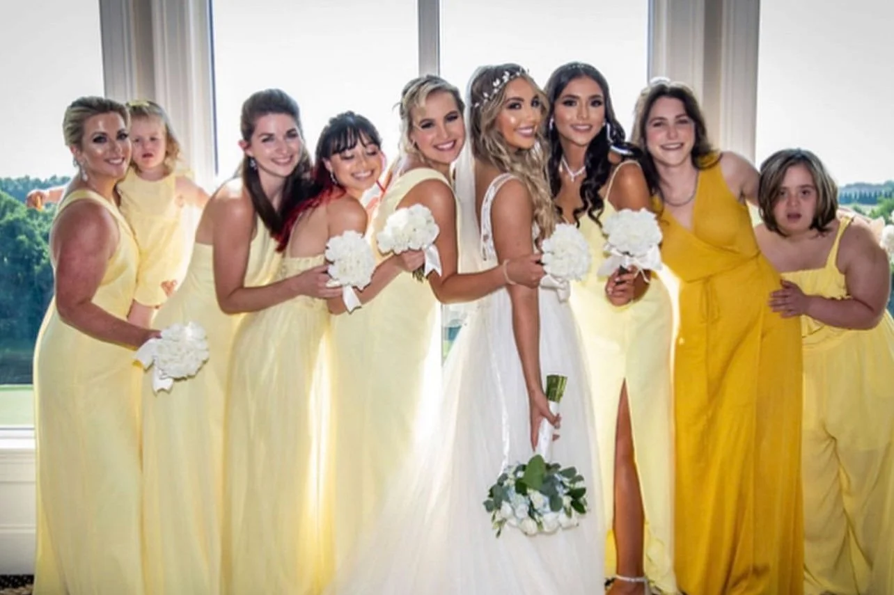 Group of women and two young girls in yellow dresses, standing together in a room with large windows, during a wedding celebration.