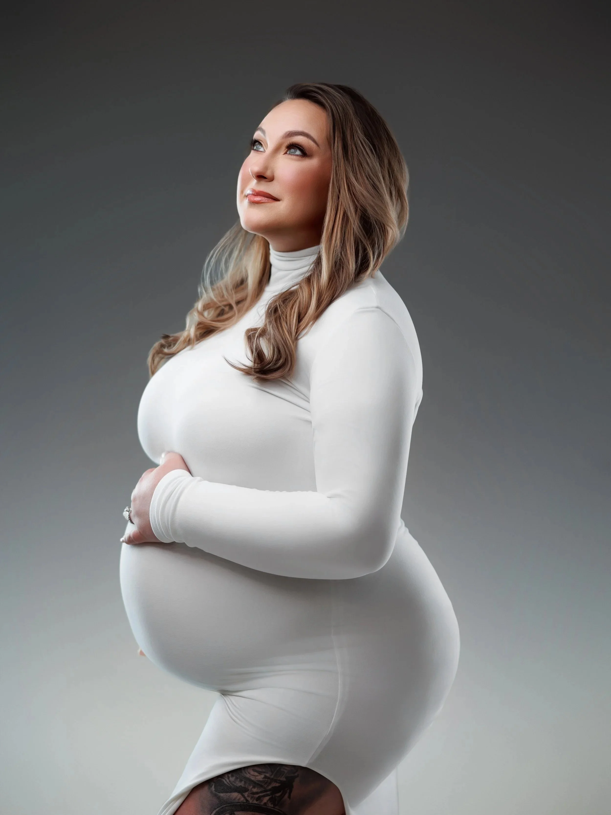 A pregnant woman with long wavy hair in a white long-sleeve dress, looking thoughtfully to the side, standing against a gray gradient background.