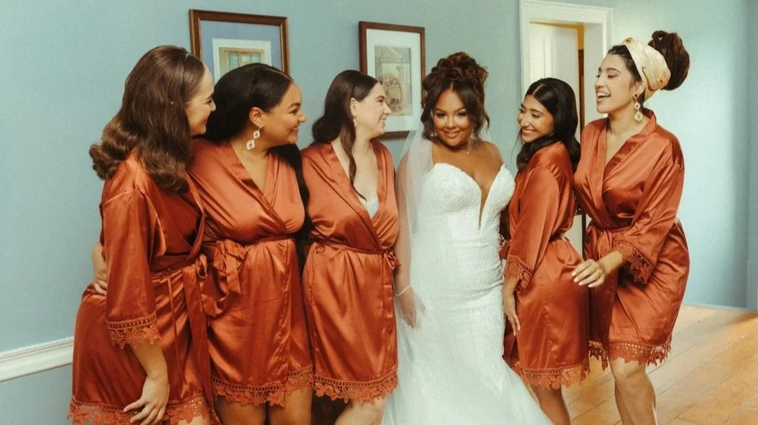 Bridal party with the bride in a white wedding gown and five women in matching rust-colored satin robes, standing together in a room with framed pictures on the wall.