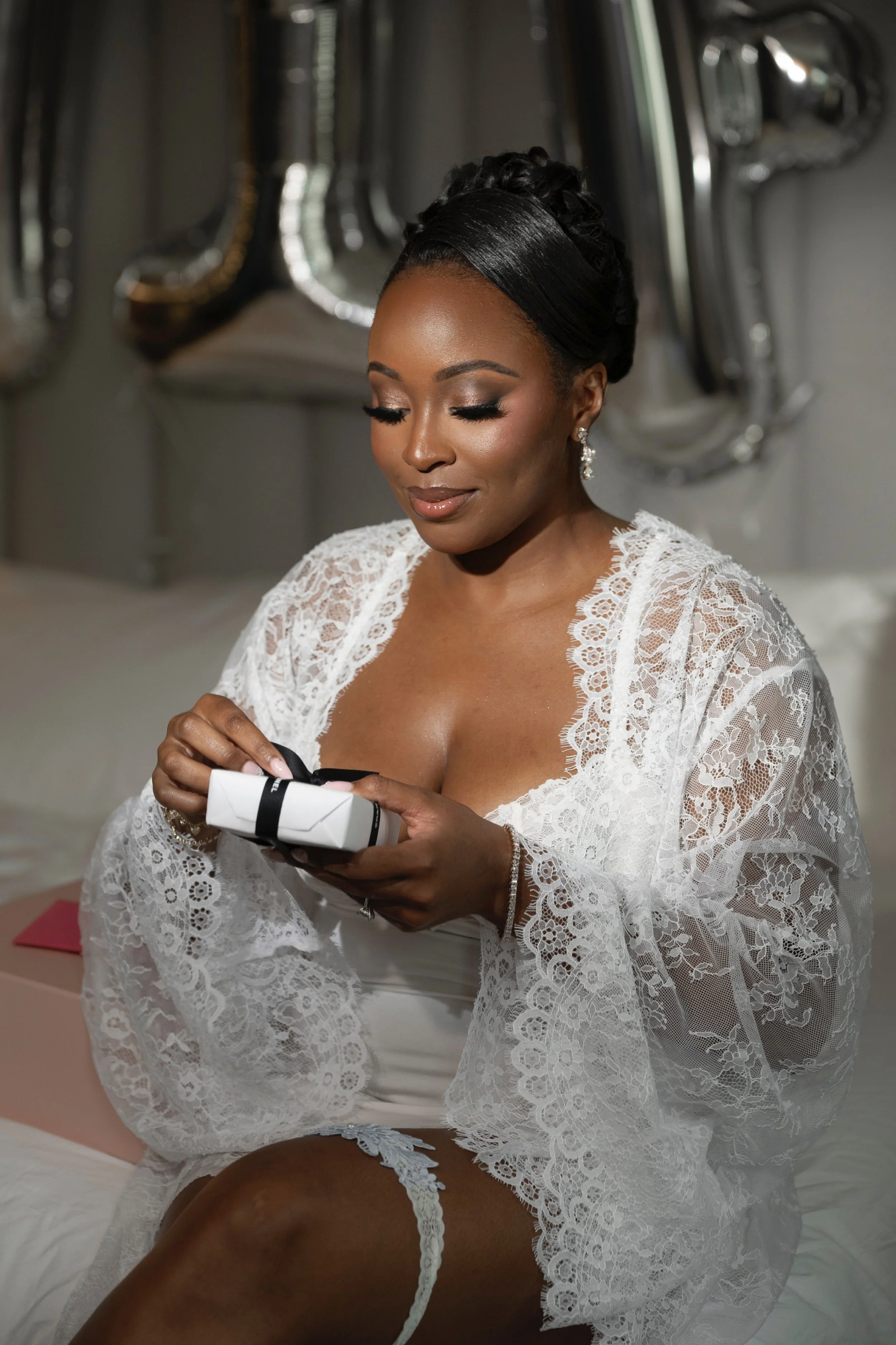 Brunette woman in wedding lace robe holding a small gift box with a black ribbon, sitting on a bed with silver 'LOVE' balloon in the background.
