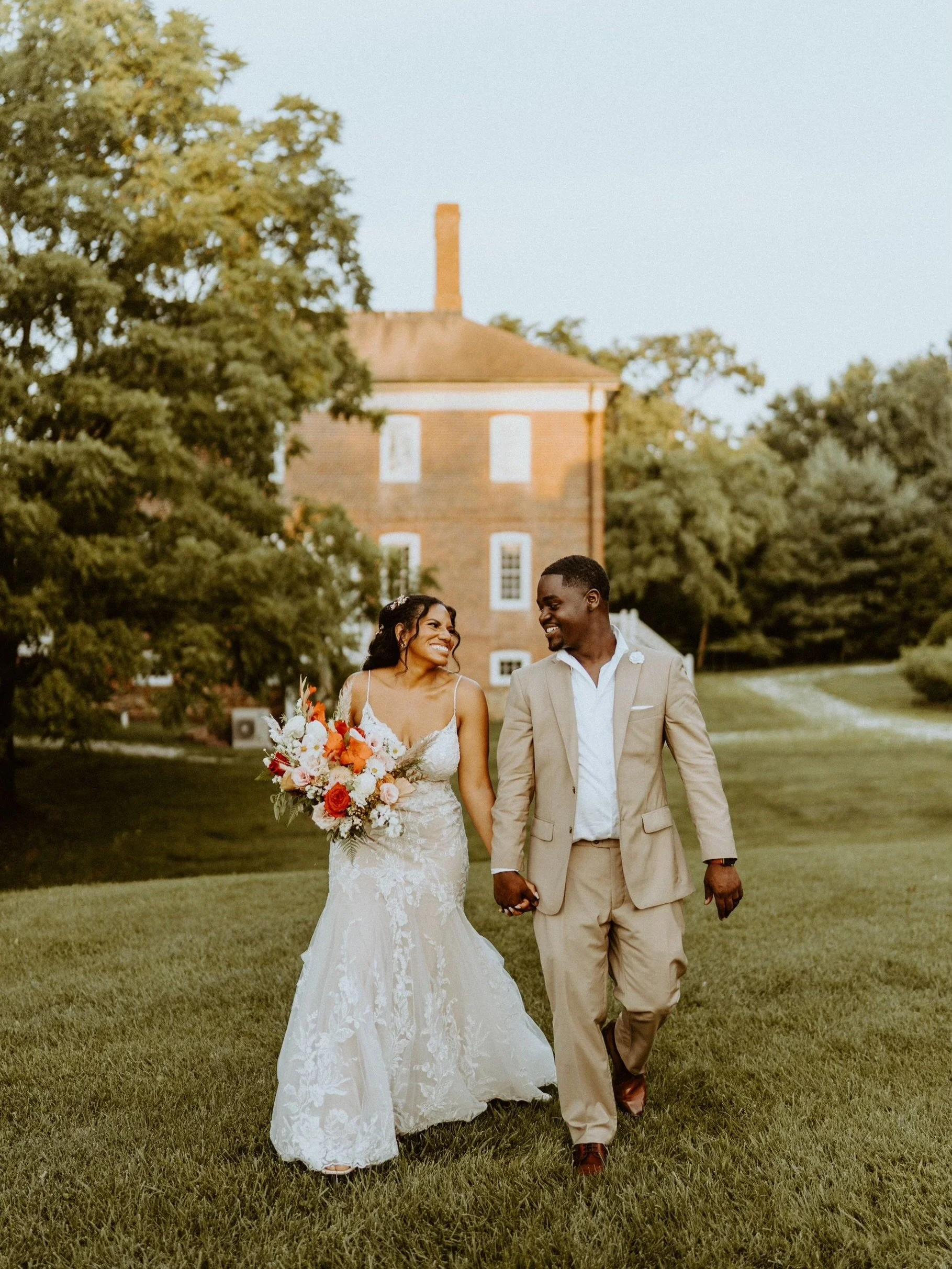 A couple in wedding attire walking hand in hand on a lush green lawn, smiling at each other, with a large brick house and trees in the background during the daytime.