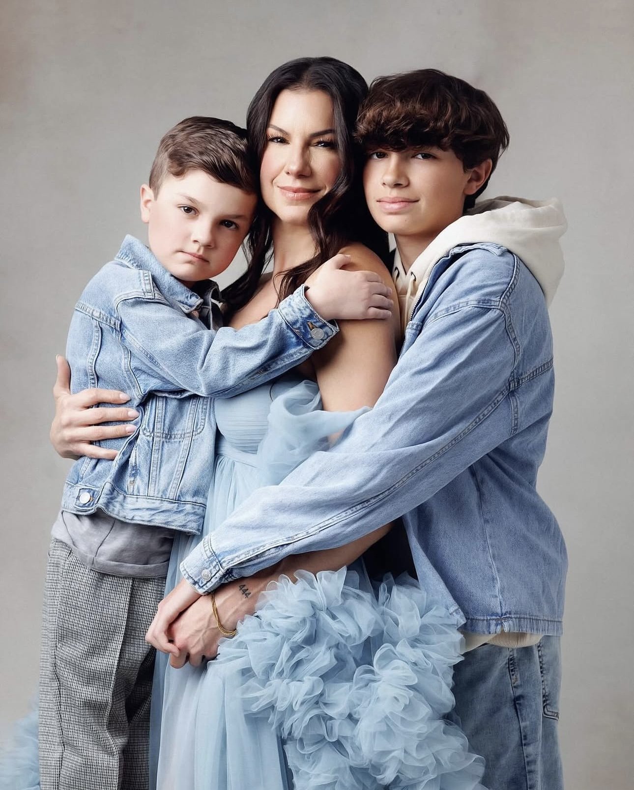 A woman and two boys wearing denim jackets hugging and smiling at the camera against a plain background.