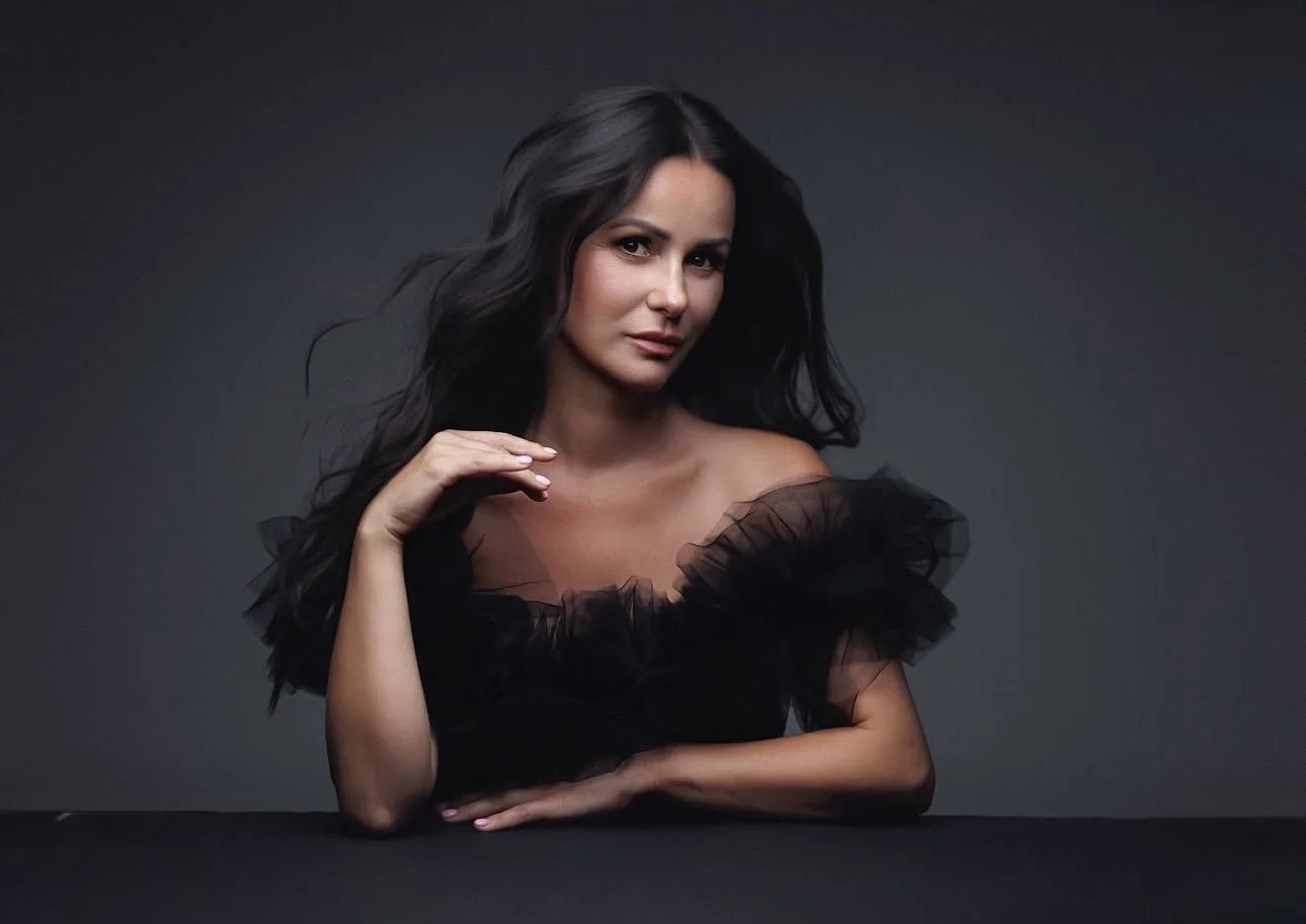 A woman with long dark hair and light skin, wearing a black tulle dress with ruffled sleeves, poses with a confident expression against a dark background.