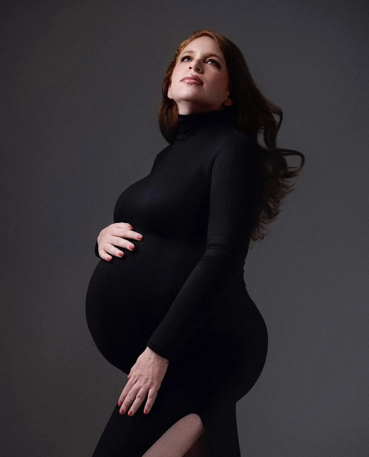 A pregnant woman with long red hair wearing a black turtleneck dress and posing against a dark gray background.