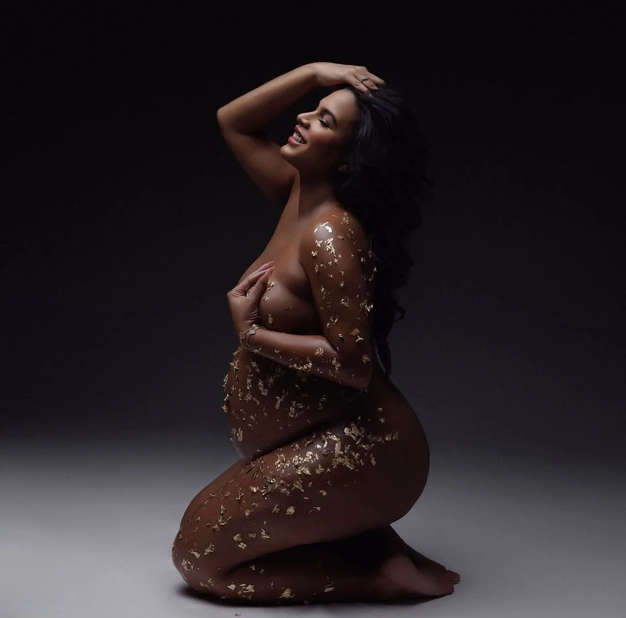 A woman with dark hair smiling, kneeling on the ground, covered in gold leaf flakes on dark background.