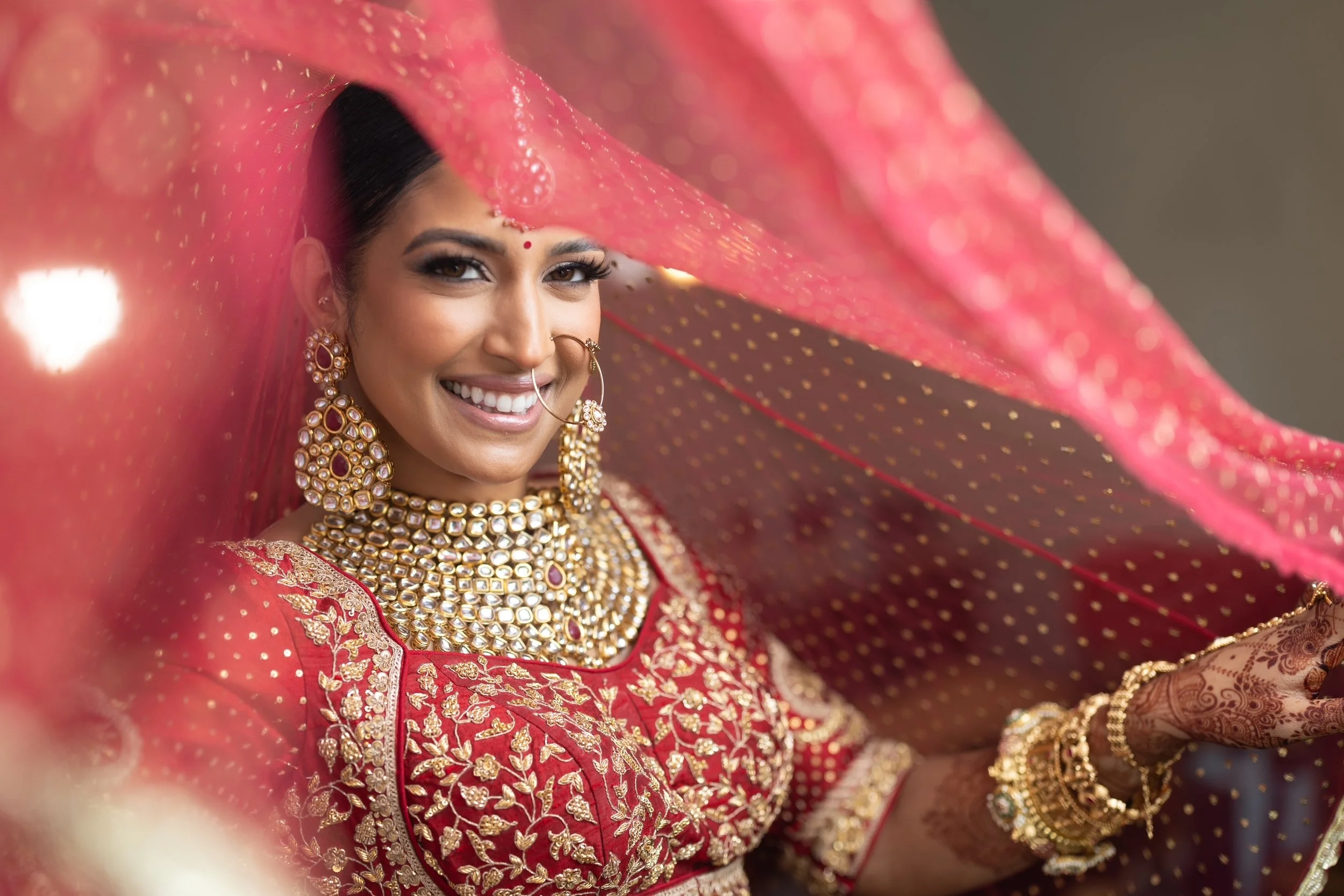 A smiling woman dressed in traditional Indian wedding attire with gold and pink jewelry, a red embroidered saree, and henna on her hands, holding a pink veil overhead.