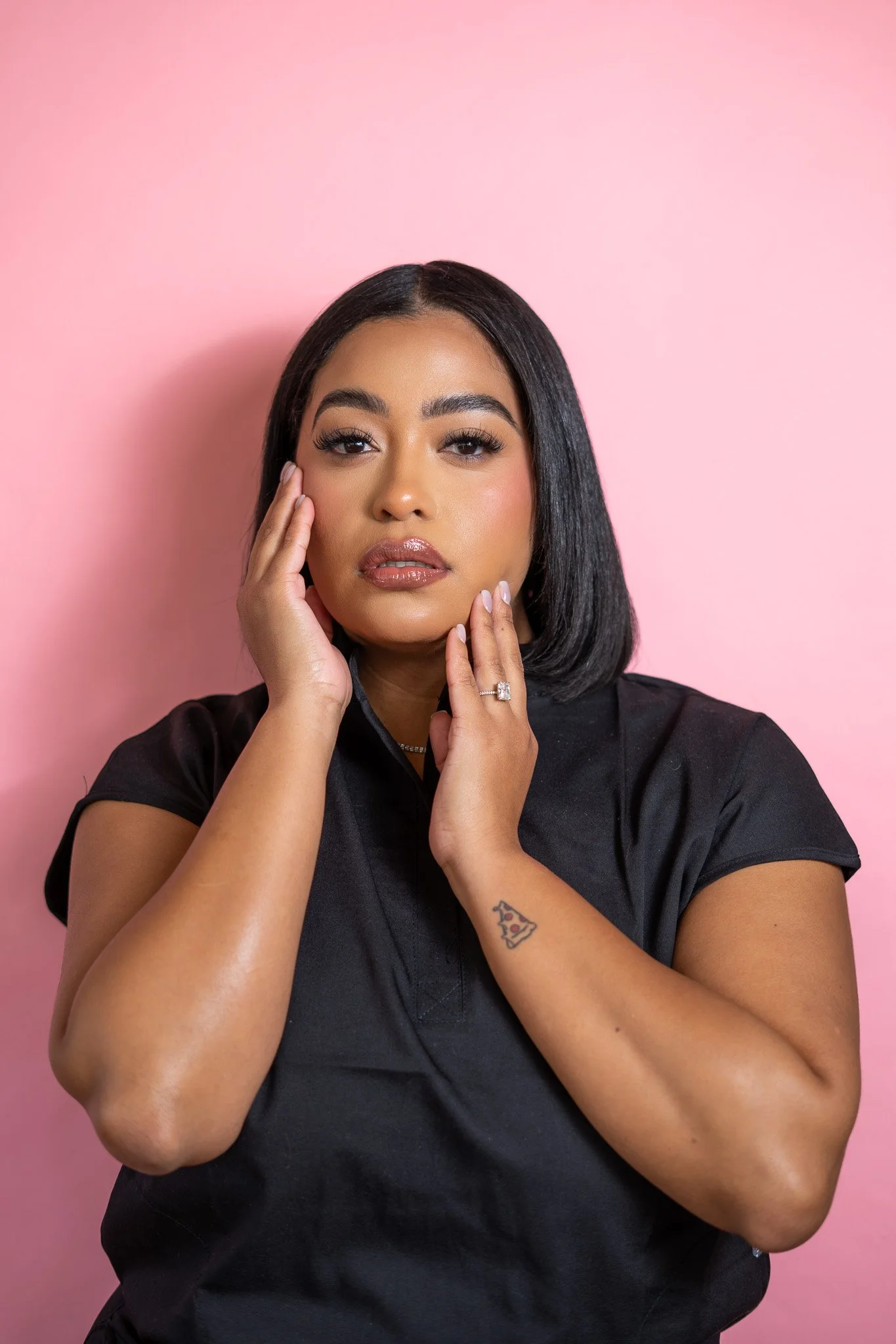 Portrait of a woman with black hair, wearing a black top, posing against a pink background.
