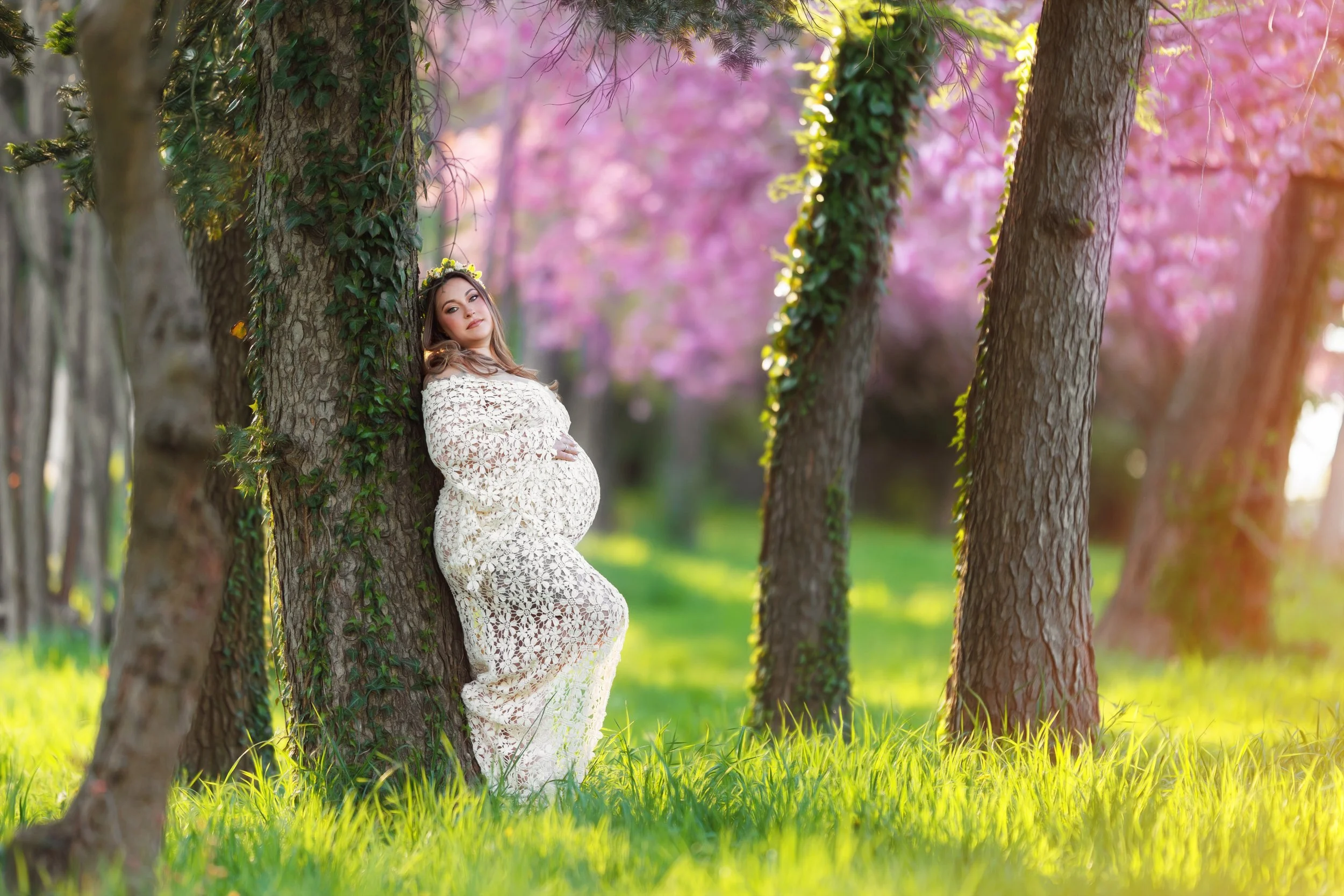 Pregnant woman in a cream white crochet dress leaning against a tree in a park with green grass and pink flowering trees in the background