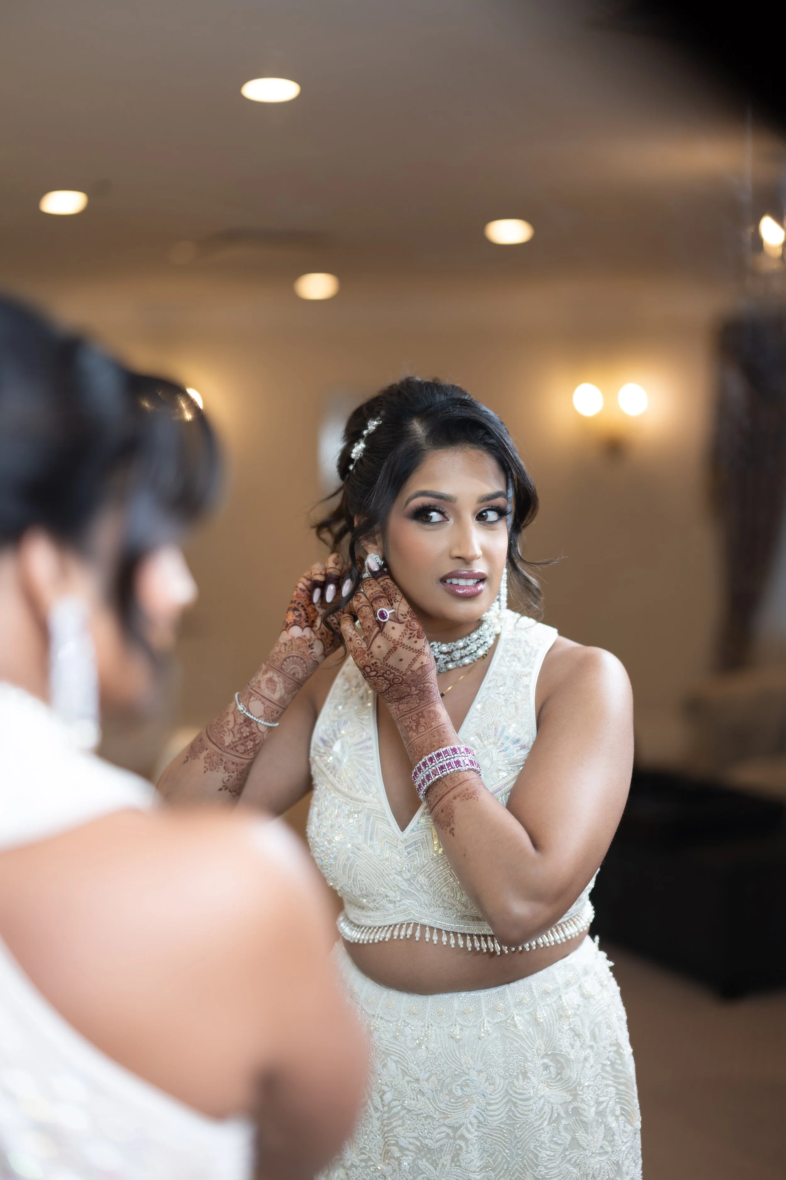 A woman dressed in traditional Indian attire, putting on earrings, with henna on her hands and jewelry, preparing for a celebration or wedding.