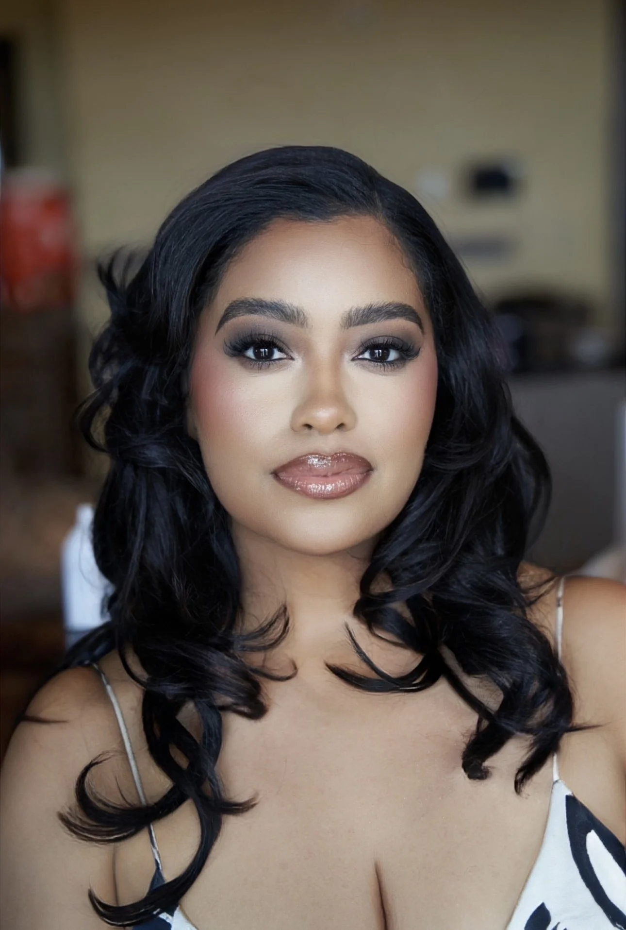 Close-up of a woman with dark, curly hair and makeup, wearing a sleeveless dress with a black and white pattern, smiling softly at the camera.