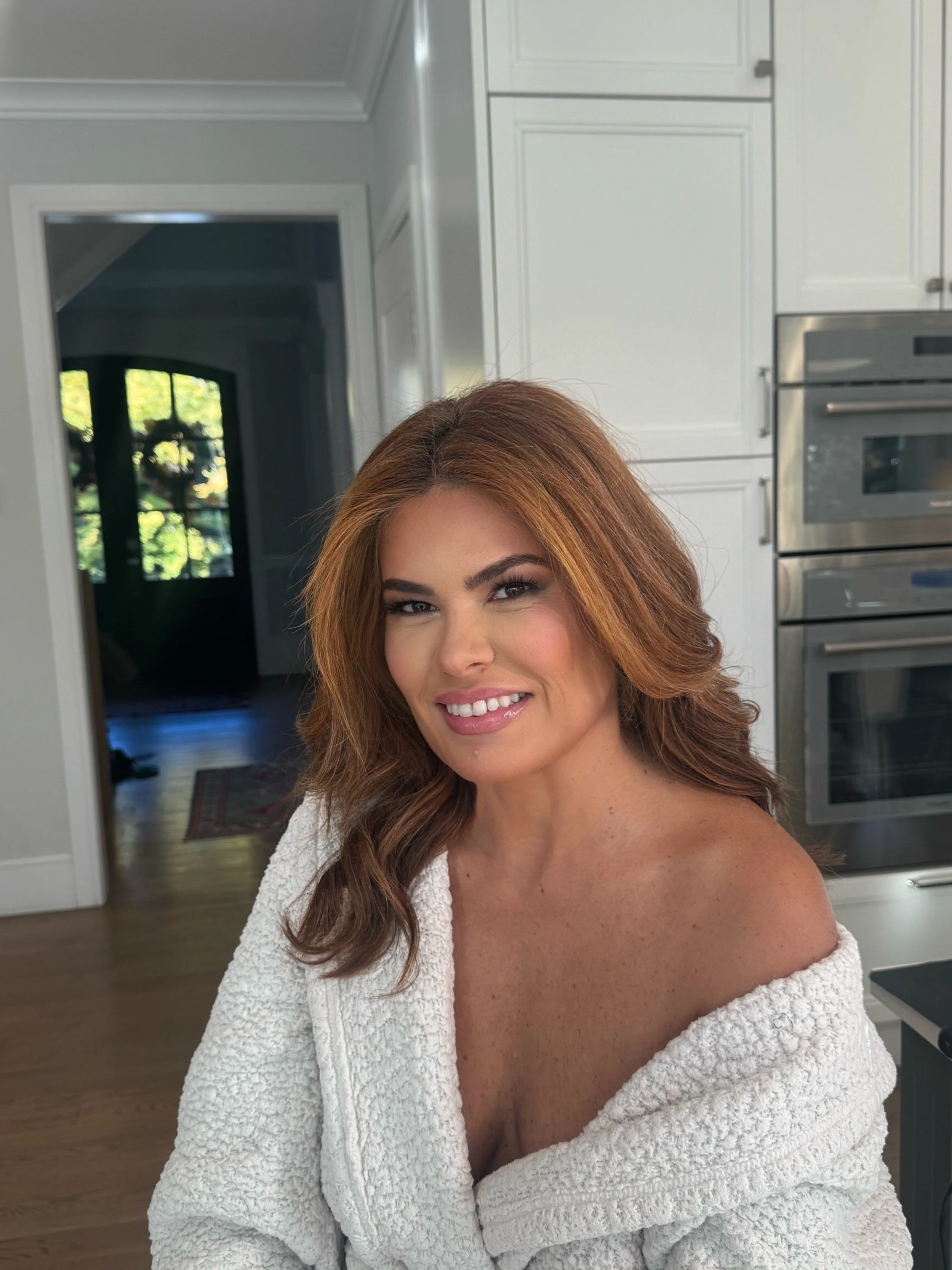 A woman with auburn hair, smiling, wearing a white bathrobe with her hair and makeup done