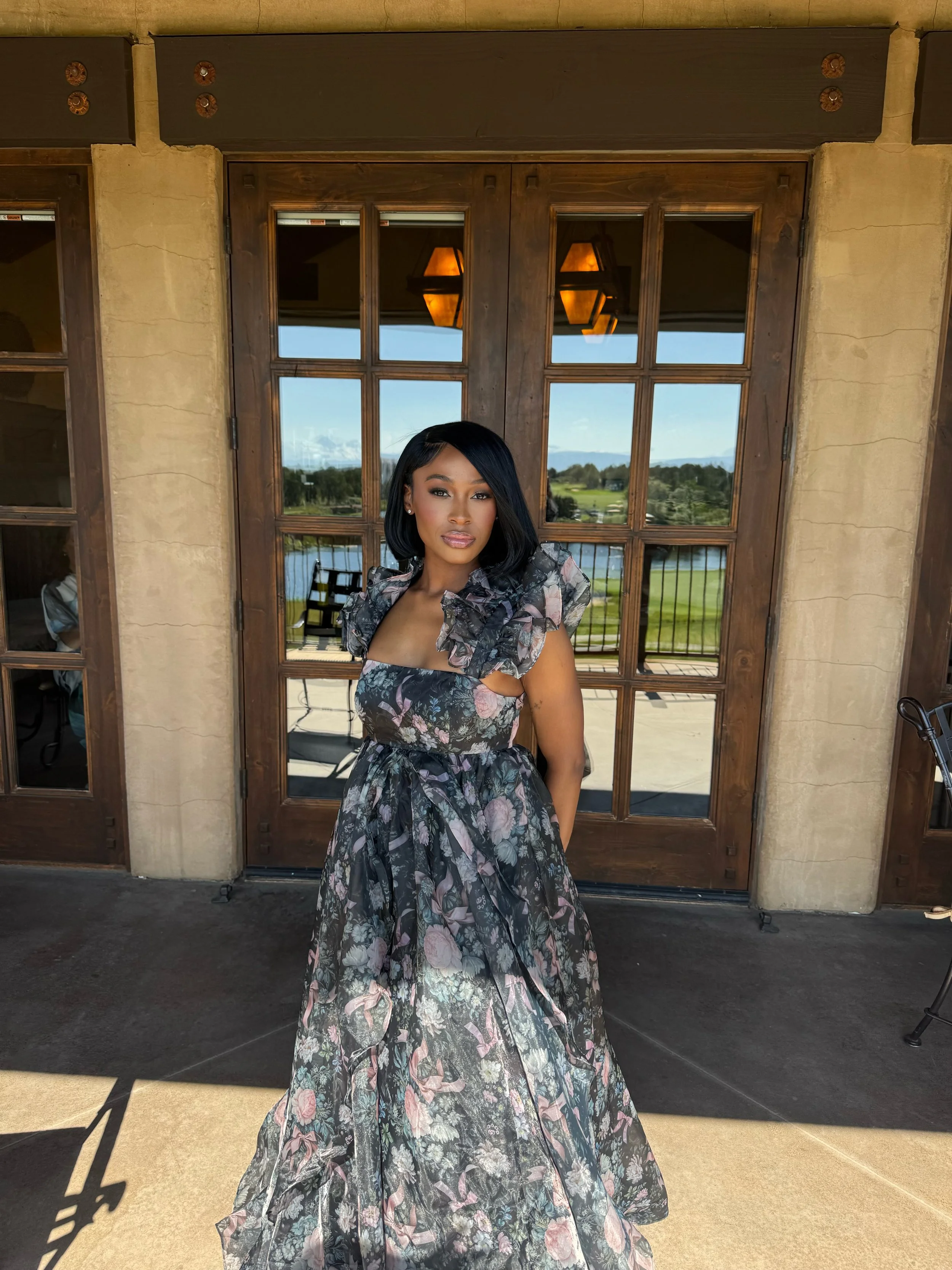 A woman in a floral dress stands in front of wooden glass doors with a view of a lake and landscape behind her.