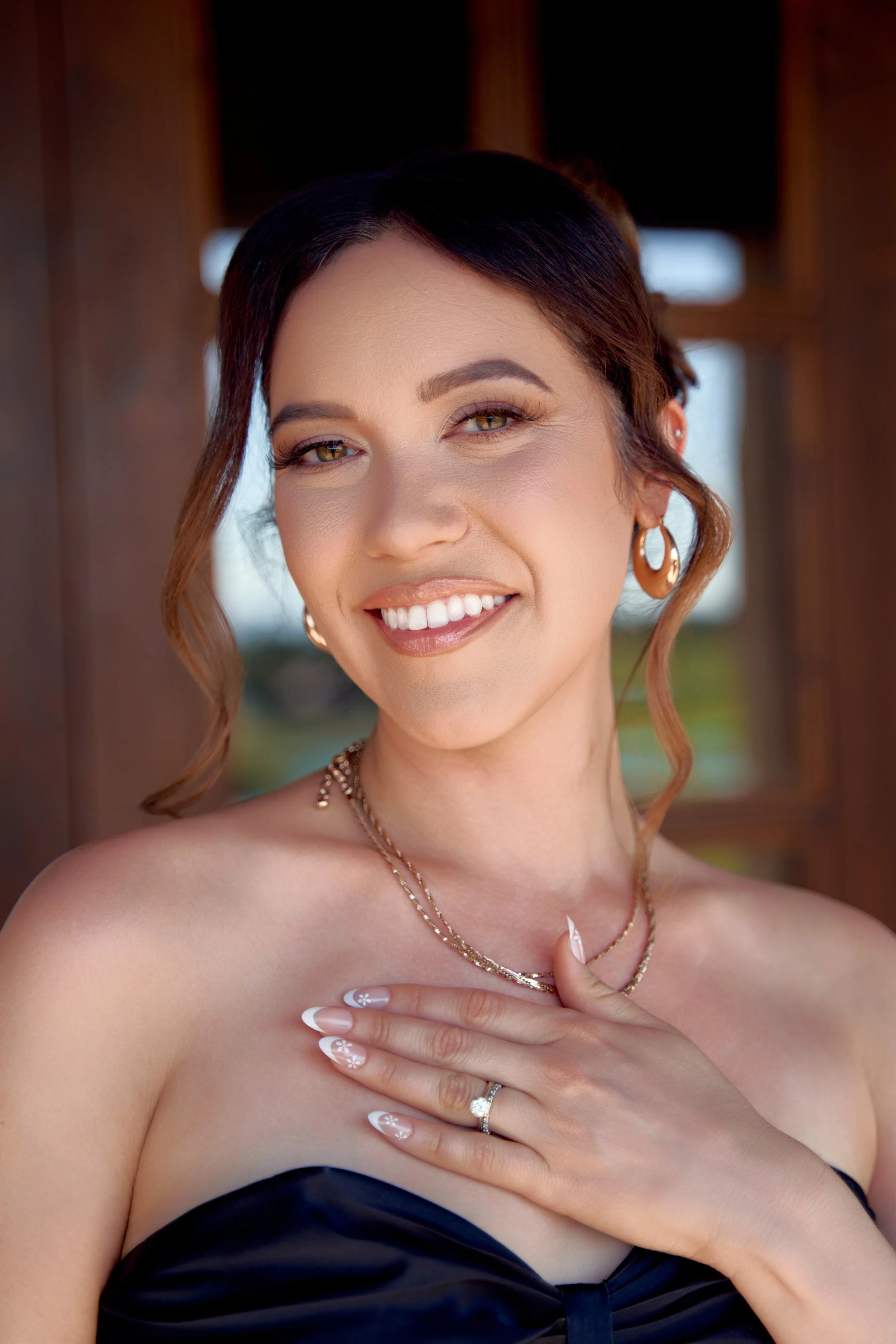 A woman smiling and looking slightly to the side, wearing earrings, layered necklaces, and a ring, with her hand on her chest, in a strapless black dress, outdoors with natural light.