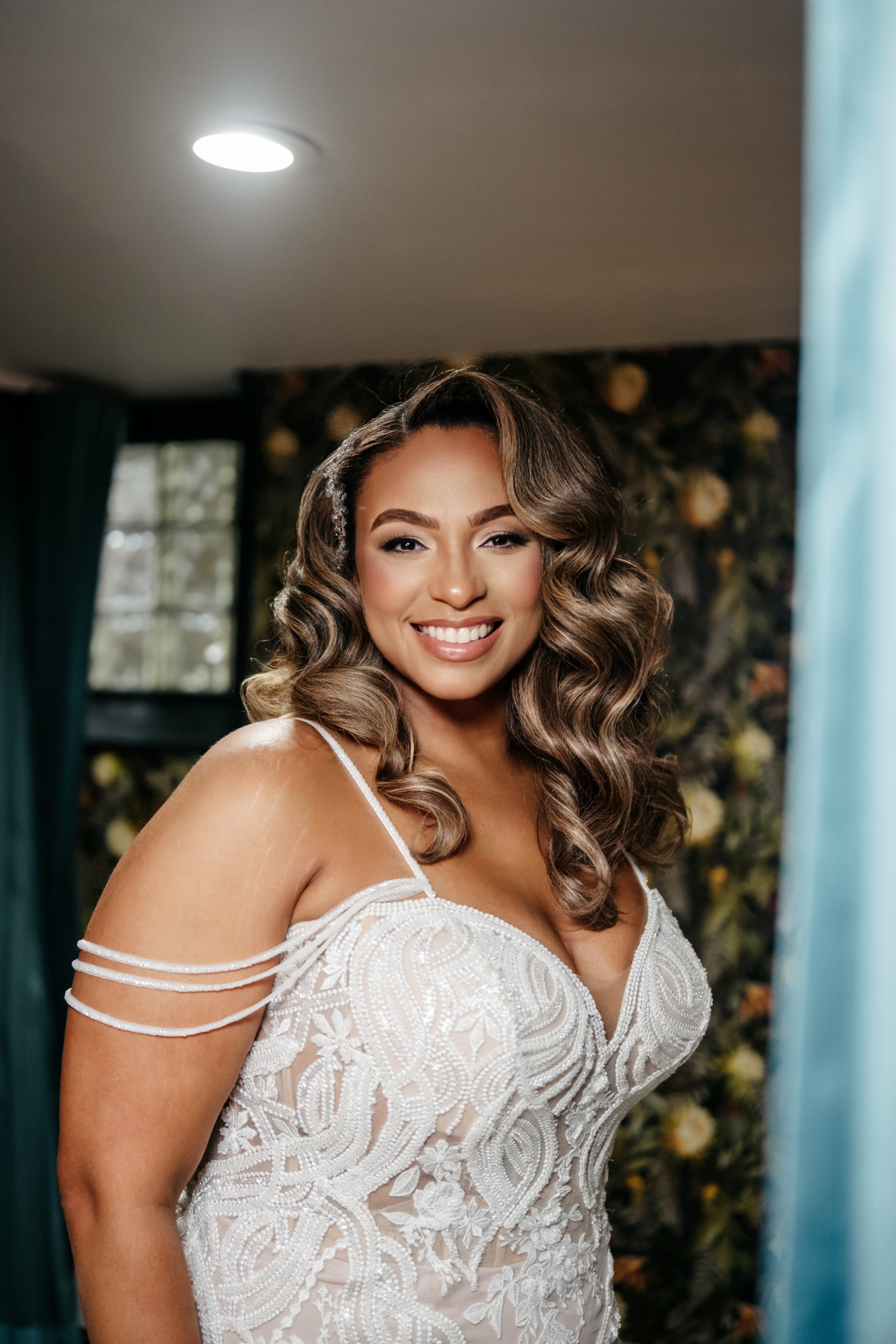 A smiling woman with wavy brown hair in a white, detailed, strap-shoulder dress standing indoors with dark floral wallpaper background and a window.