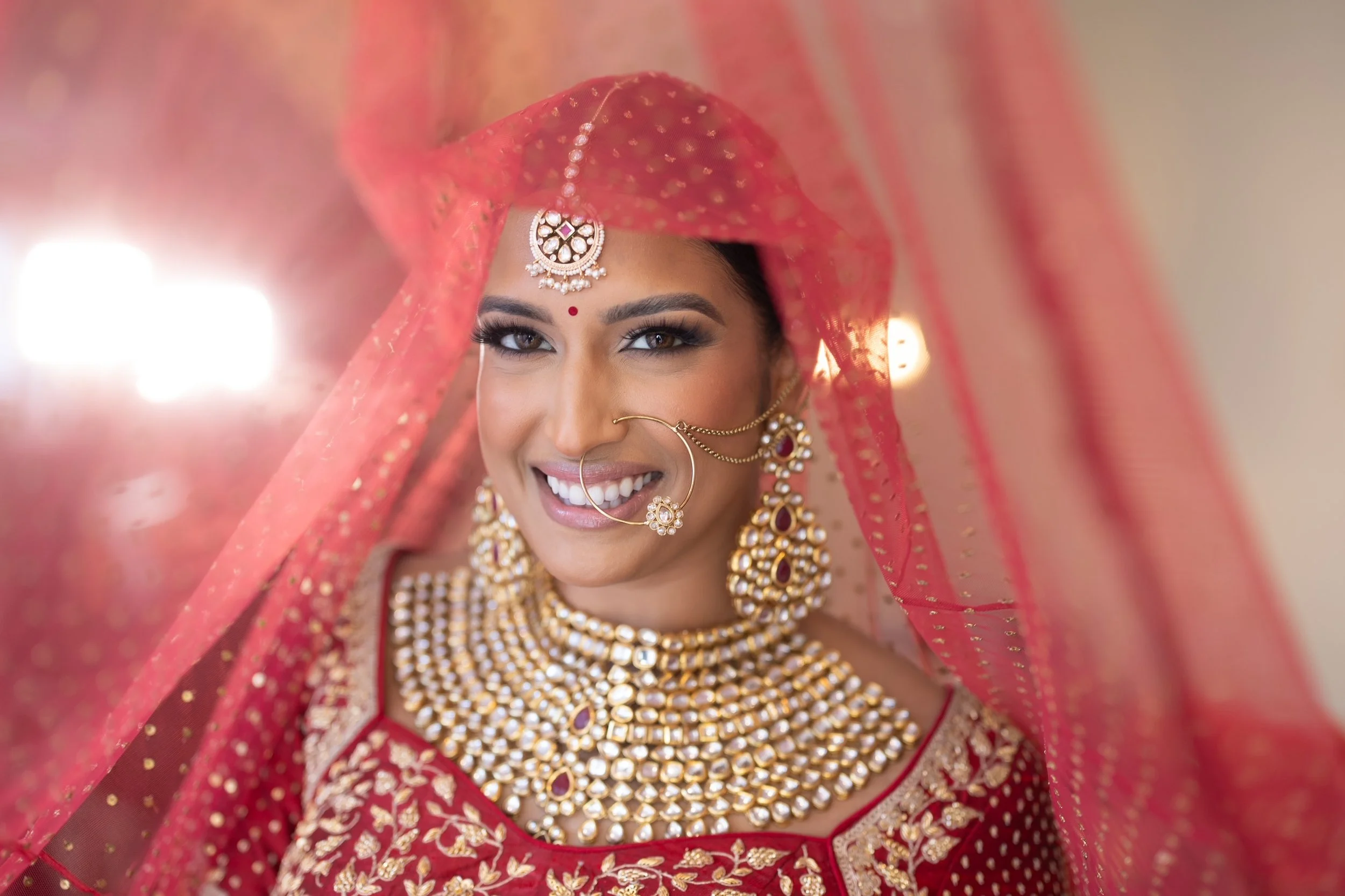 Close-up of a smiling Indian bride wearing traditional red and gold jewelry and a red bridal saree, with a sheer dupatta covering her head.