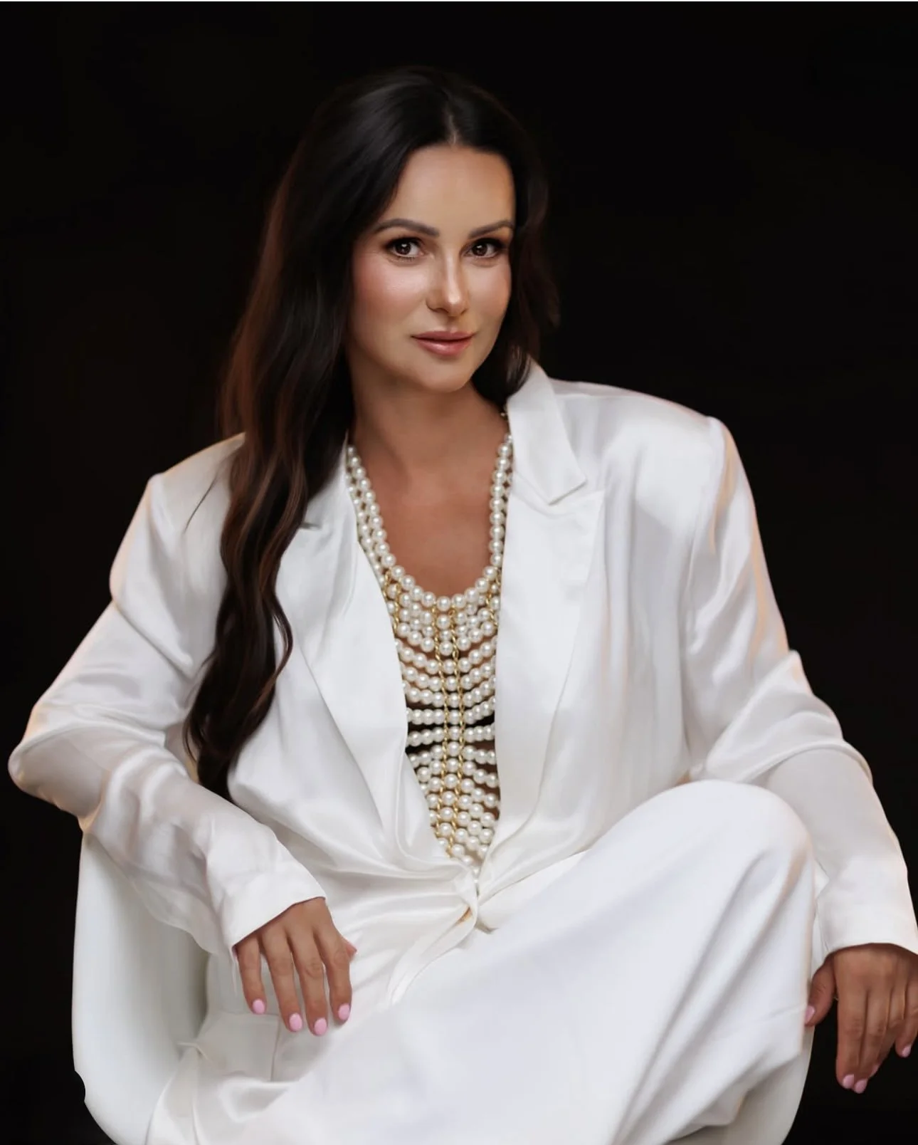 A woman with long dark hair wearing a white suit and layered pearl necklaces, sitting against a black background.