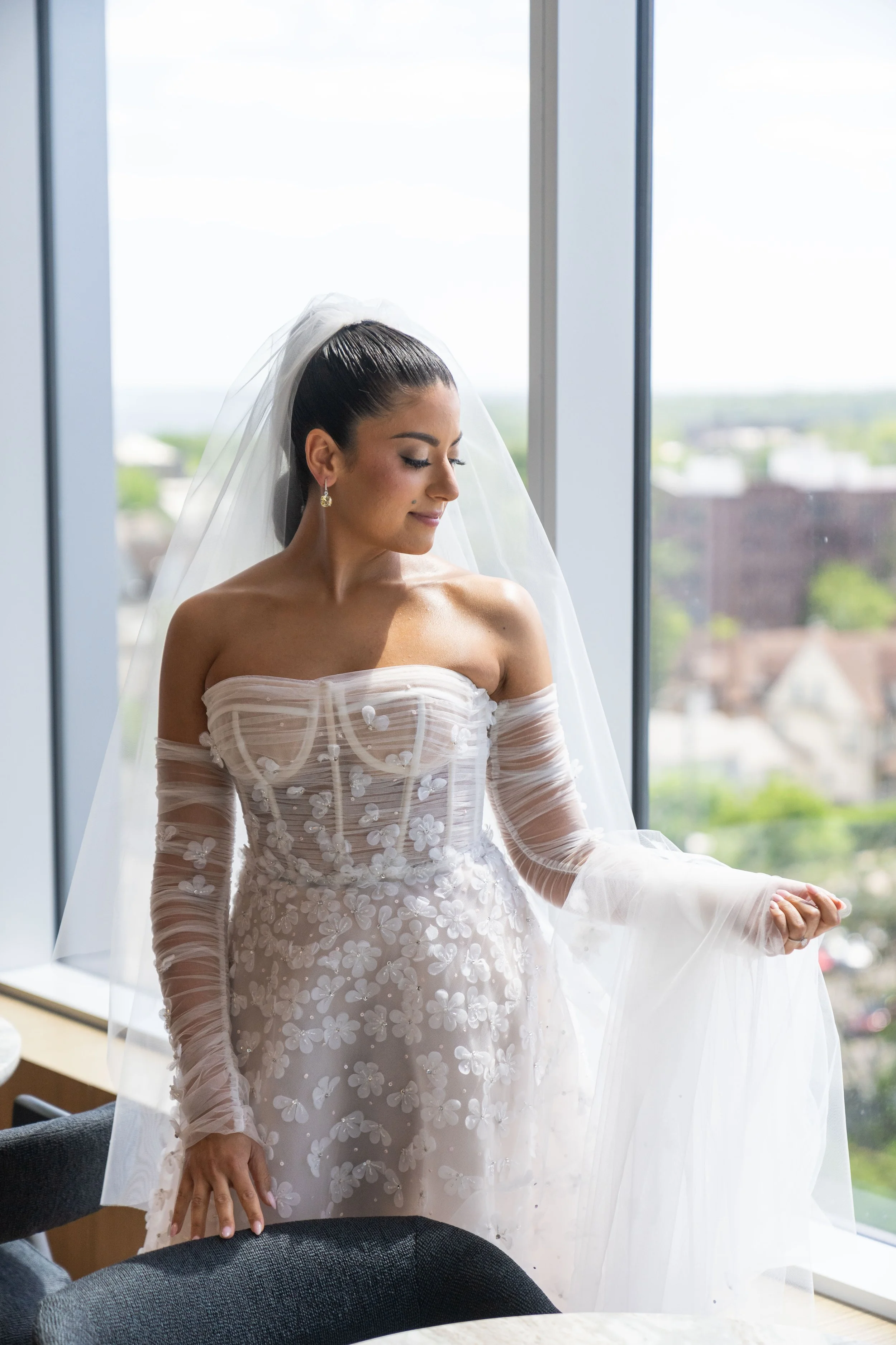 Bride in wedding dress and veil standing near large window with city view.