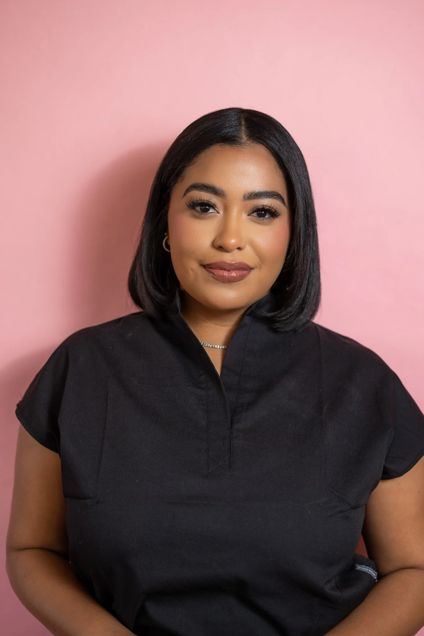 Portrait of a young woman with shoulder-length black hair, wearing a black top, against a pink background.