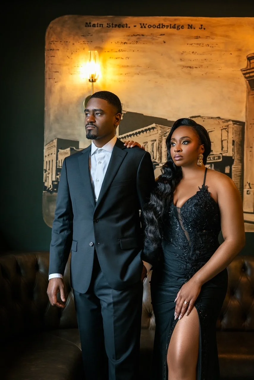 A well-dressed man in a black suit and white shirt standing next to a woman in a black gown with lace details and a high slit, inside a room with a vintage-style mural behind them and a wall-mounted light fixture.