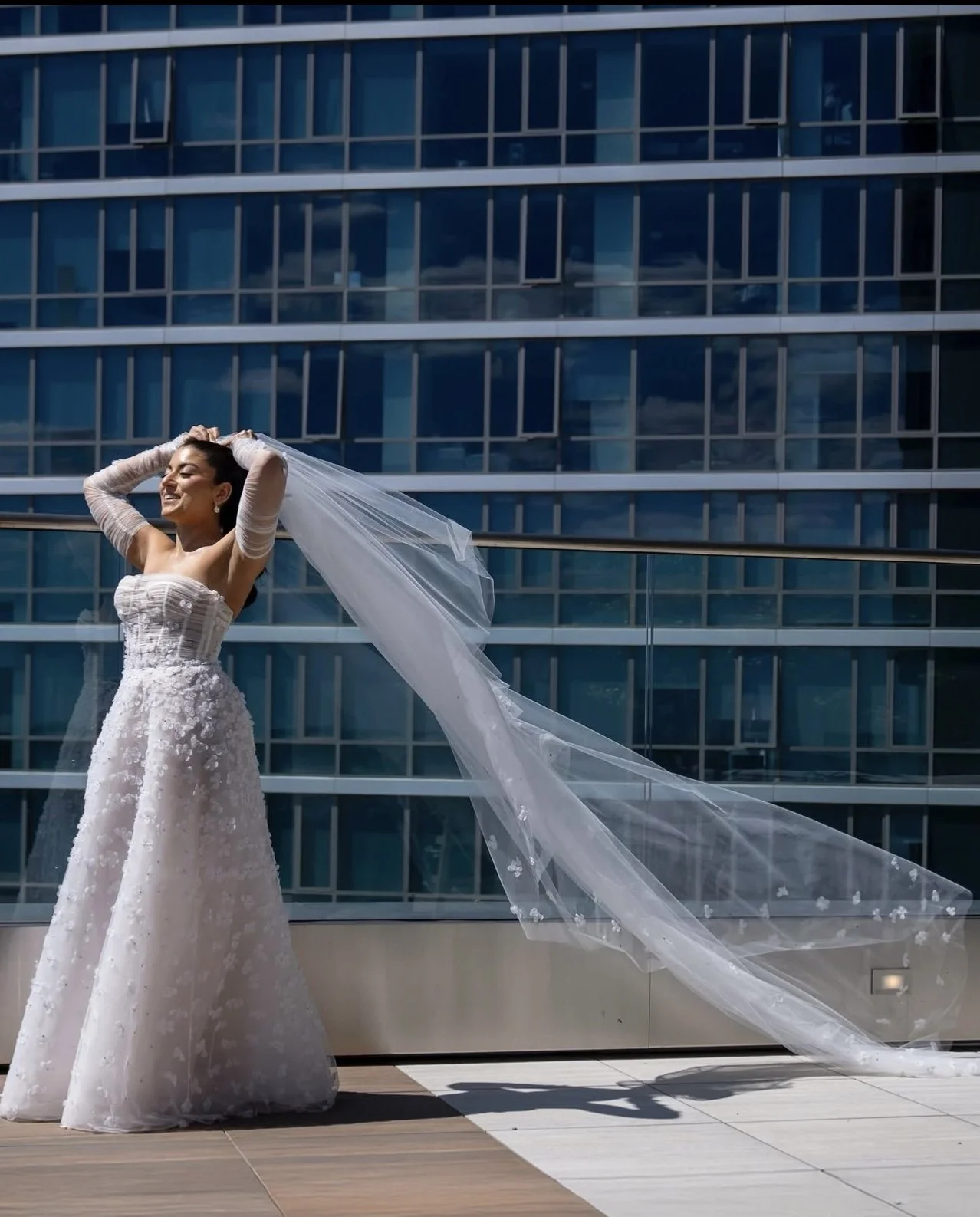A bride in a white wedding dress and veil standing outdoors in front of a modern glass building, smiling with her hands on her head and her veil flowing to the side.