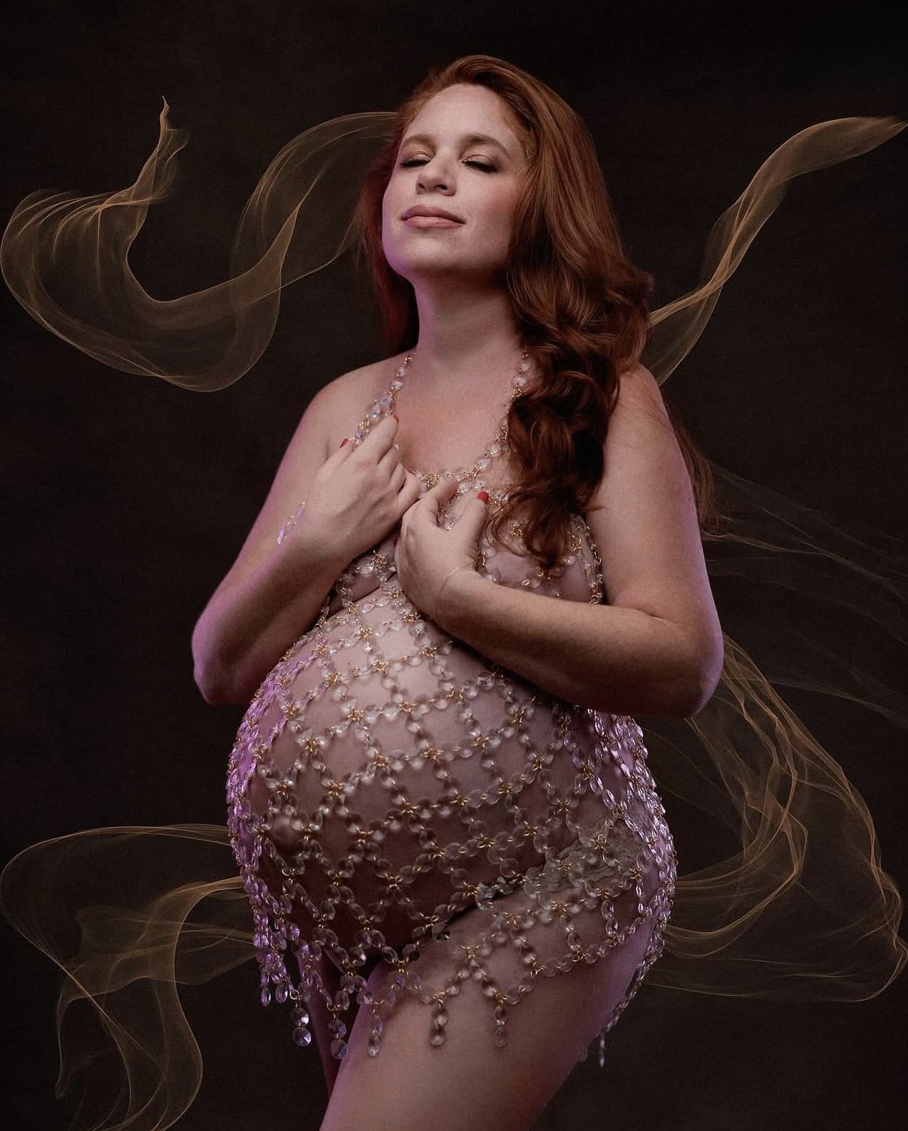 A woman with red hair wearing a jewelry dress, eyes closed, with a dark background and light effects around her.