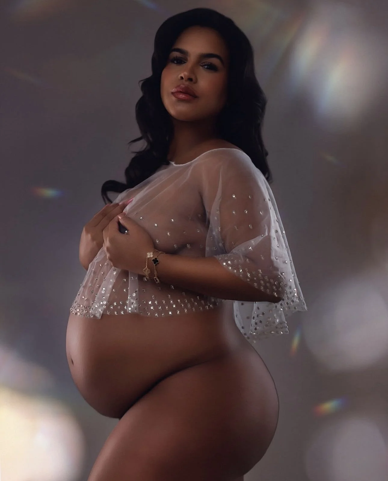A pregnant woman with long dark hair, wearing a sheer, embellished top, holding the top close to her chest, with her hand resting on her abdomen. She is posing against a dark background with soft lighting and multicolored light flares.