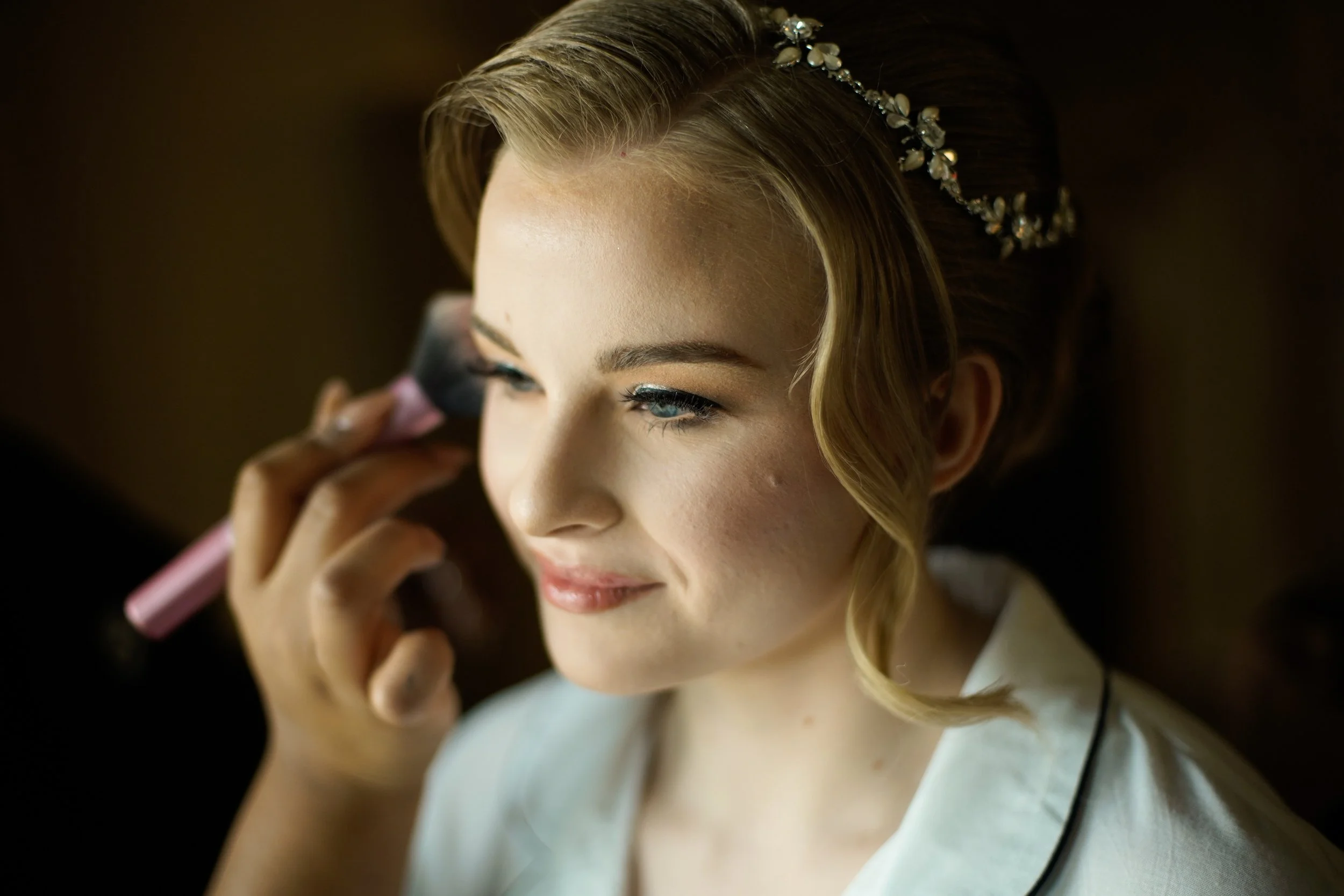 A woman with blonde hair styled in loose waves, wearing a decorative headband, has her makeup applied by a makeup artist using a pink blush brush.