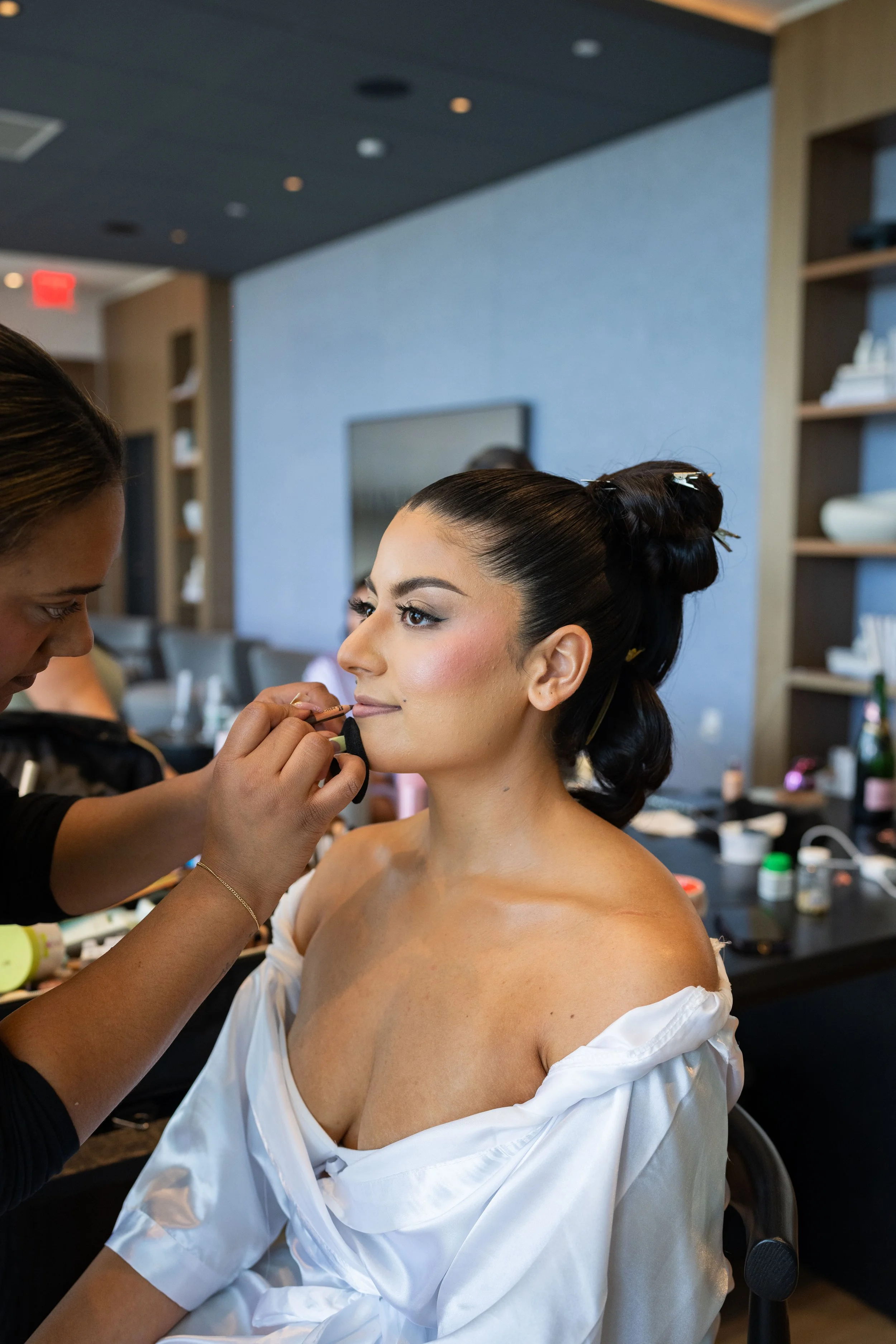 A woman with dark hair in an elegant updo is getting her makeup done by a makeup artist in a modern room.