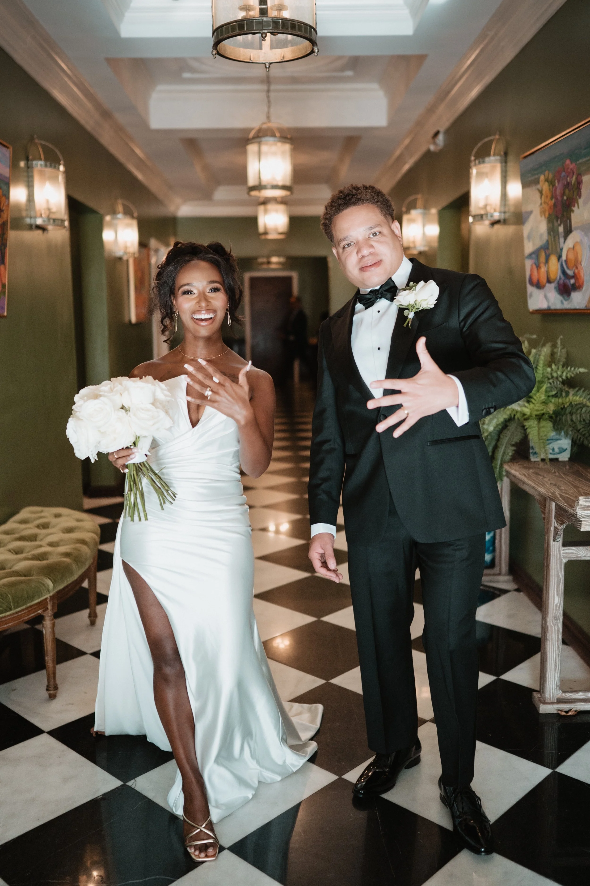 A bride and groom in wedding attire standing in a hallway with black and white checkered flooring. The bride is holding a bouquet of white roses and has her hand on her chest, smiling. The groom is dressed in a black tuxedo with a bow tie, pointing a