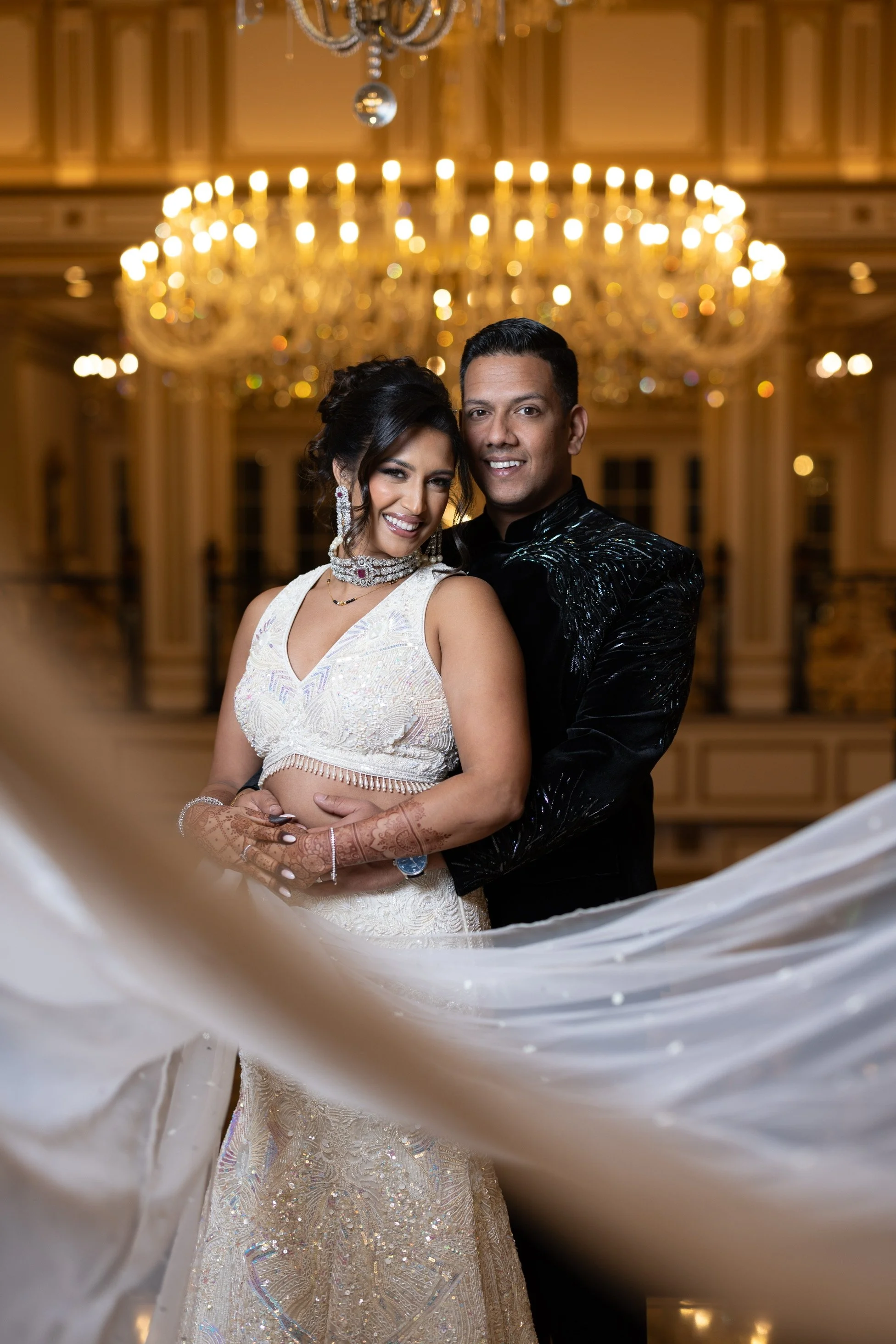 A couple in traditional wedding attire posing in an elegant, chandelier-lit ballroom.