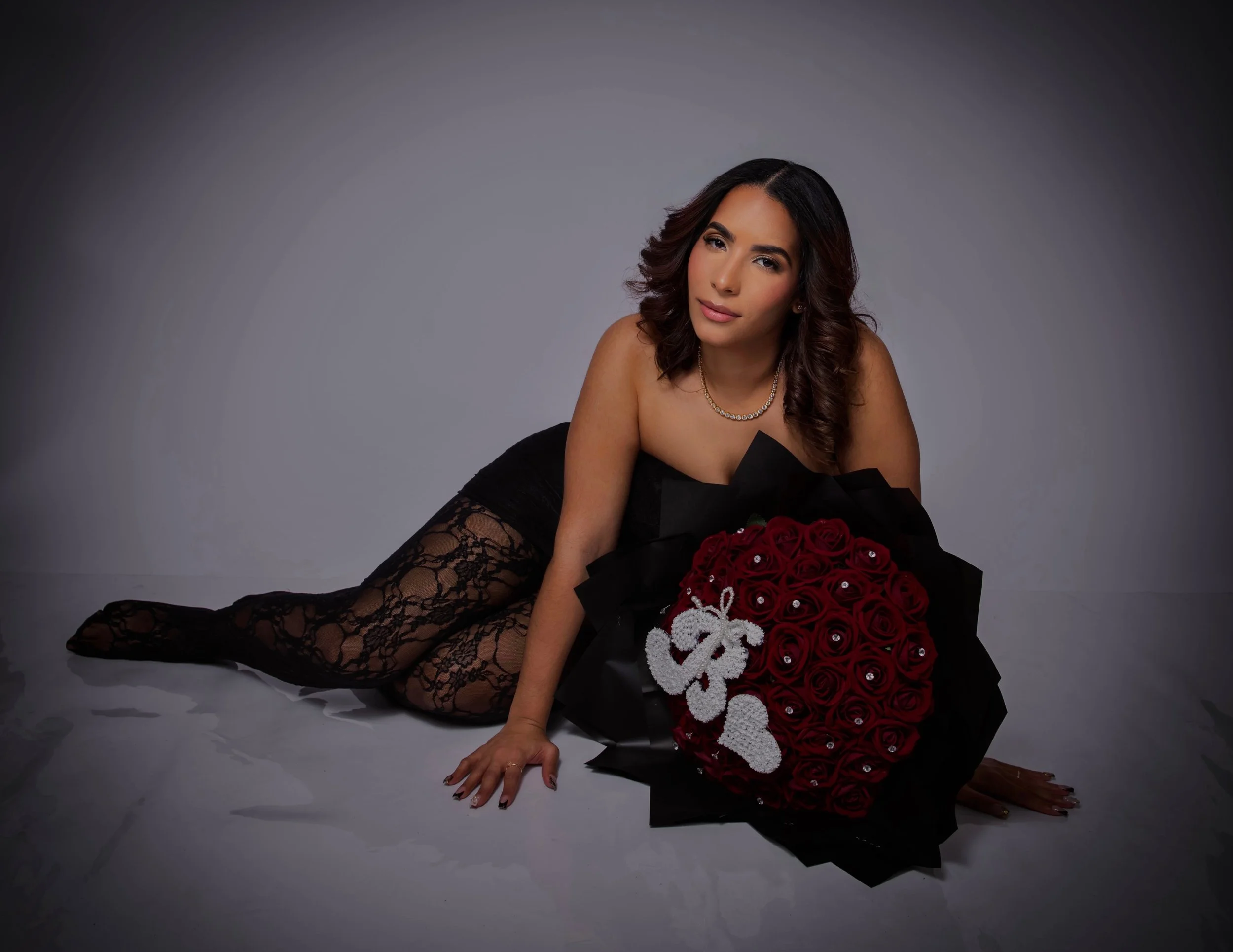 A woman with wavy dark hair lies on her side on a plain gray background, wearing black lace leggings and no top, holding a bouquet of red roses decorated with a white ribbon and the letters 'B' and 'R' in rhinestones.