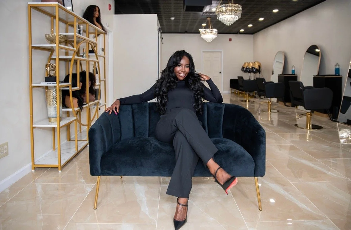 A woman in black clothing and heels sitting on a dark blue velvet couch in a modern salon with white walls, chandeliers, and black chairs.