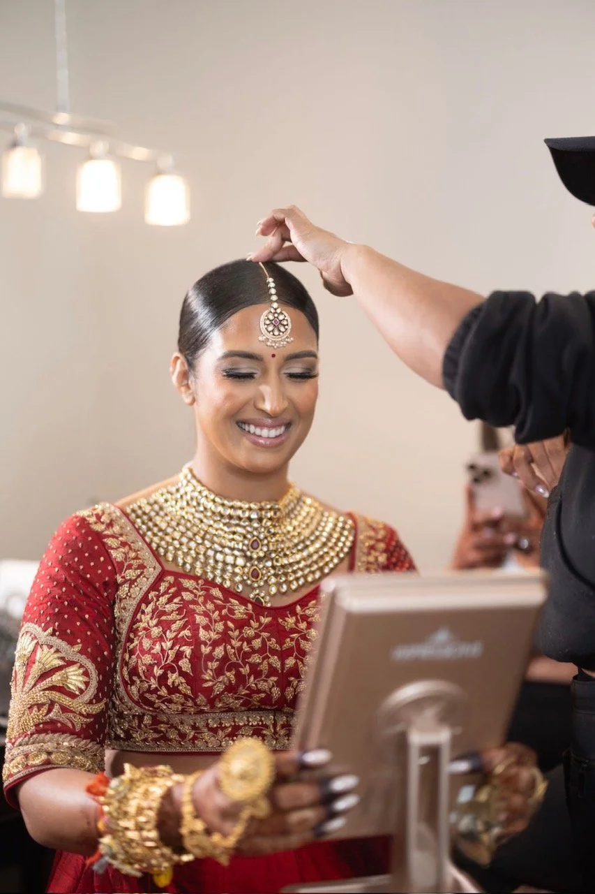 A woman in traditional Indian attire with jewelry, smiling as someone places a decorative forehead ornament on her head during a pre-wedding or festive ceremony.