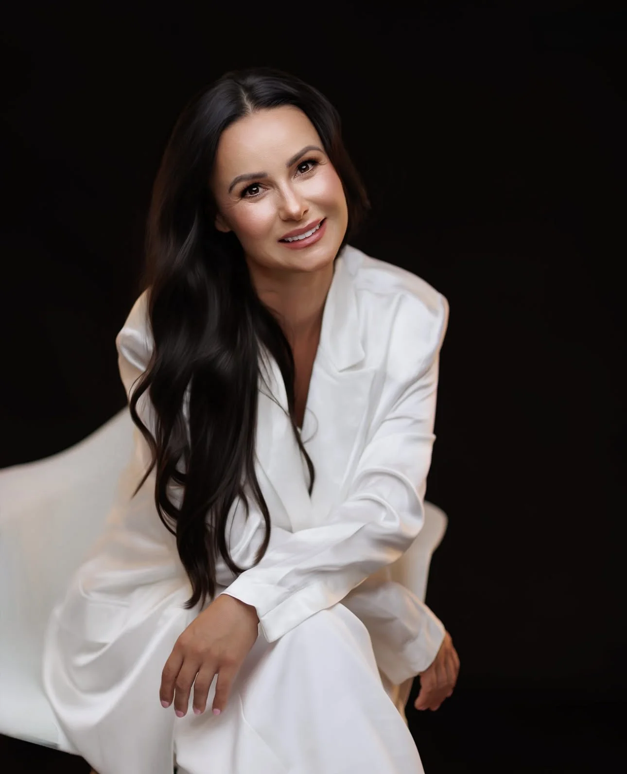 A woman with long dark hair, wearing a white satin robe, sitting on a white cushioned chair against a black background, smiling and looking at the camera.